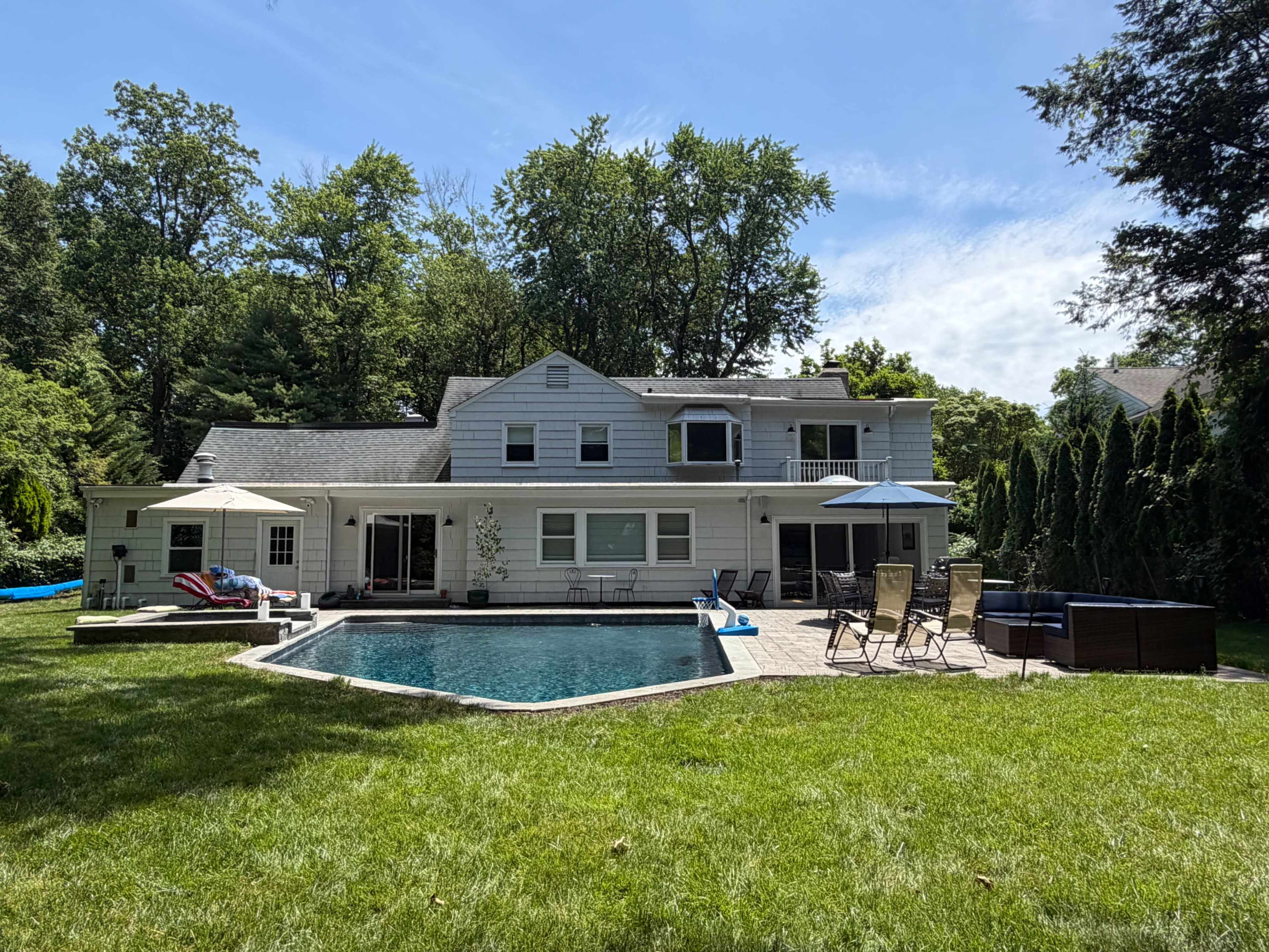 A two-story house with a pool and patio space is set against a backdrop of trees in a lush green backyard.