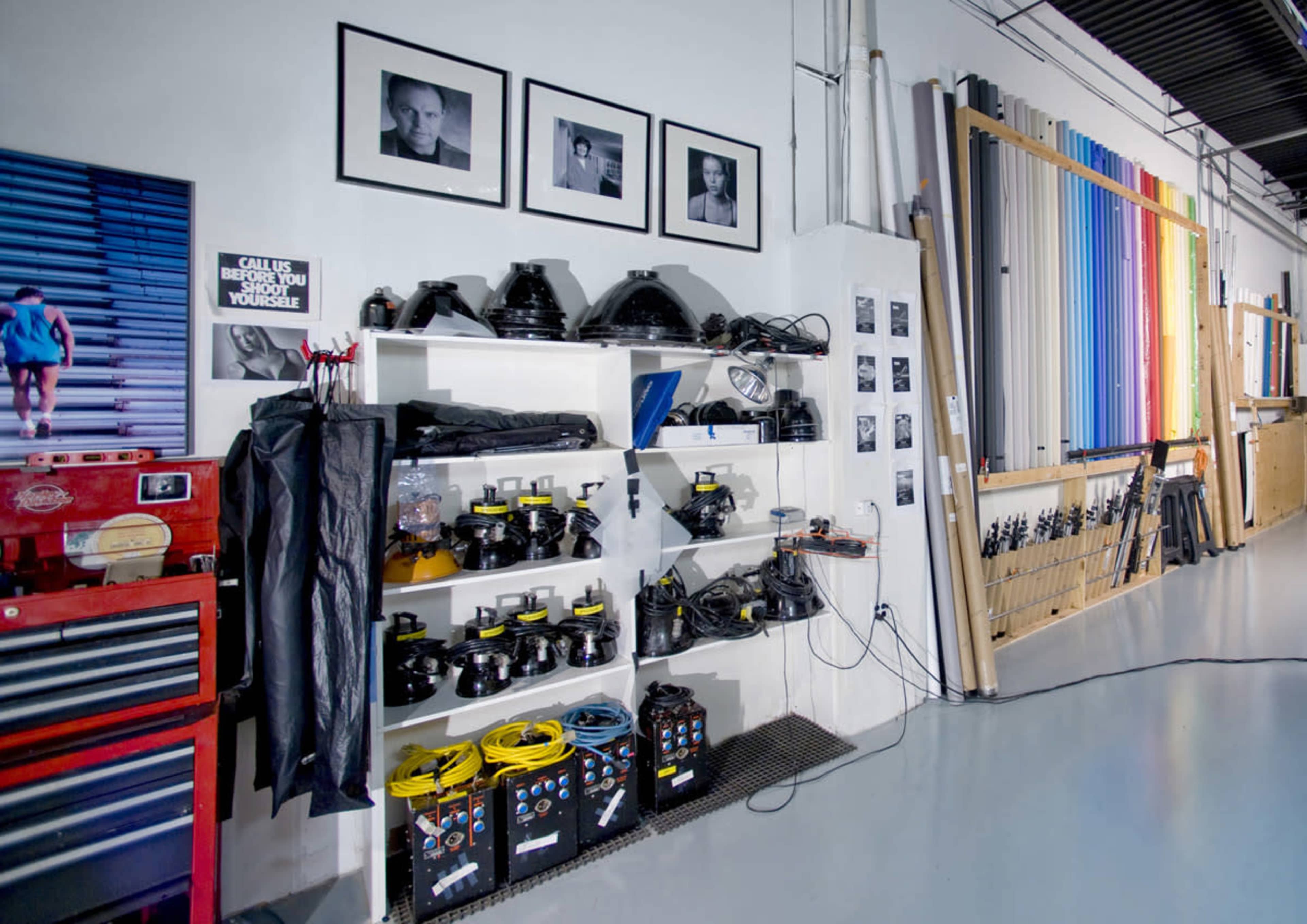 A photography studio with a shelf of equipment, including lights and cables, along with framed black-and-white portraits on the wall.