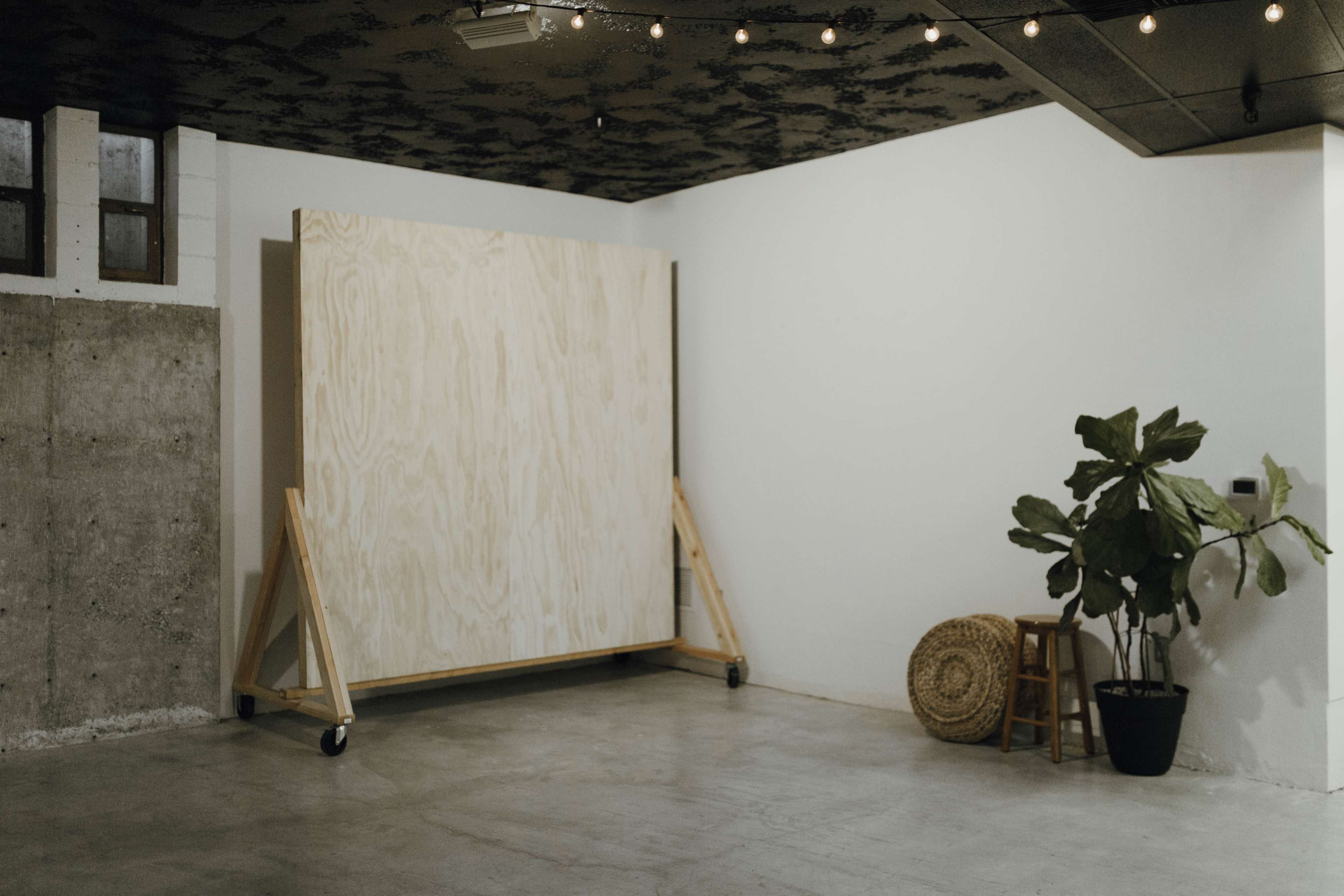 A large wooden panel stands on wheels against a bare wall in a minimalistic room, accompanied by a potted plant and a woven stool.