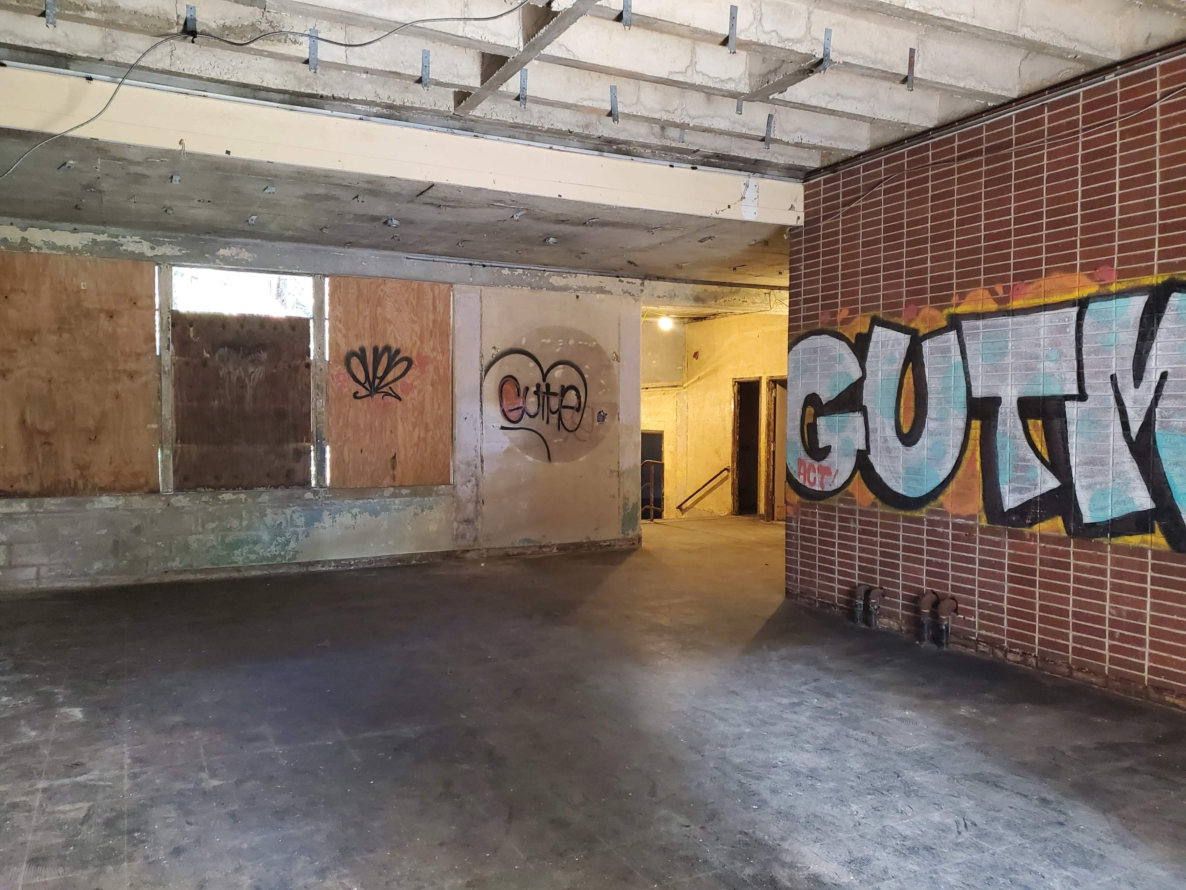 An empty, dimly lit interior space with exposed concrete walls, wooden boards covering windows, and graffiti on one wall.