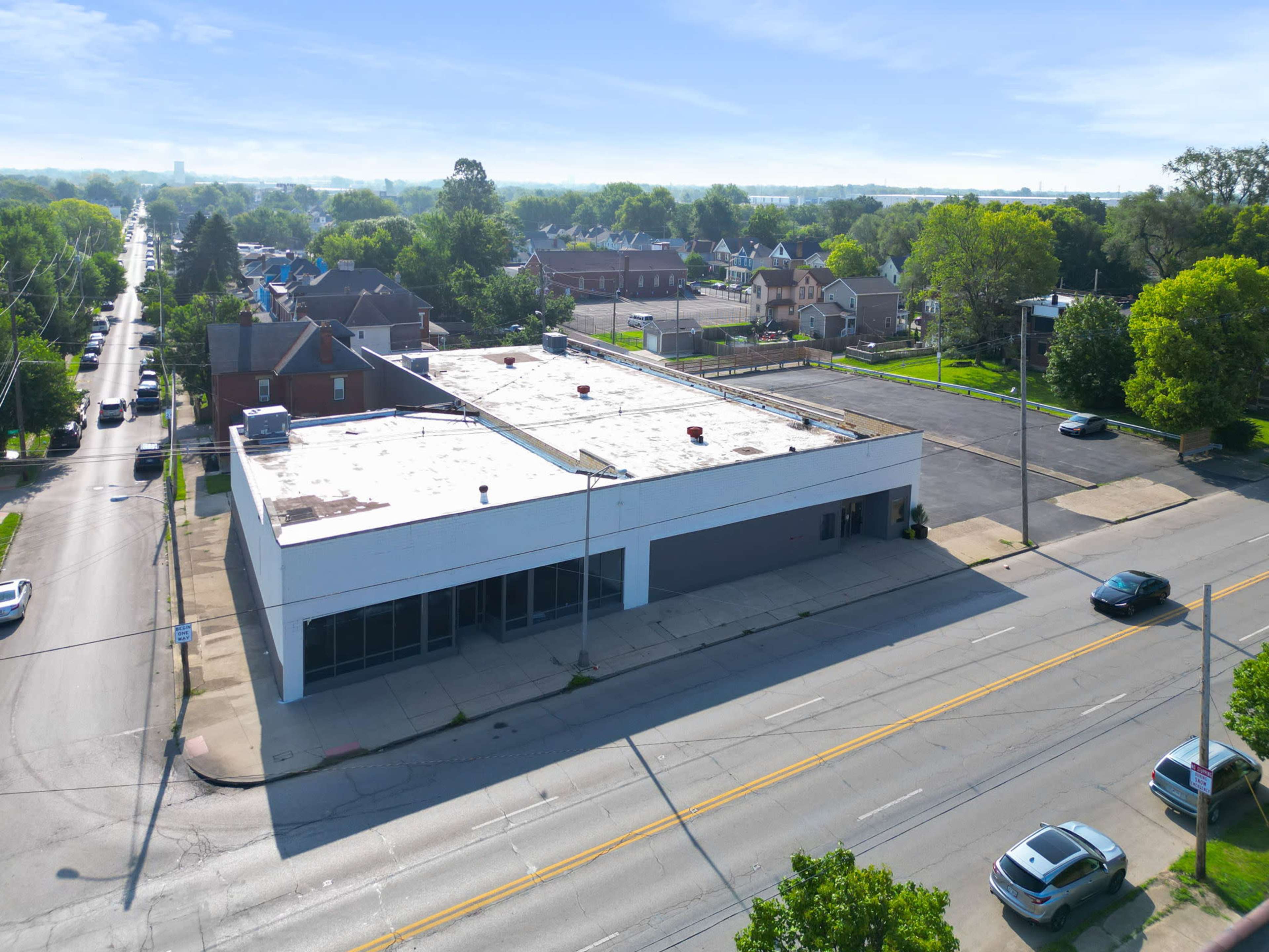 An aerial view shows a vacant commercial building at the corner of a street, surrounded by residential homes and a parking lot.