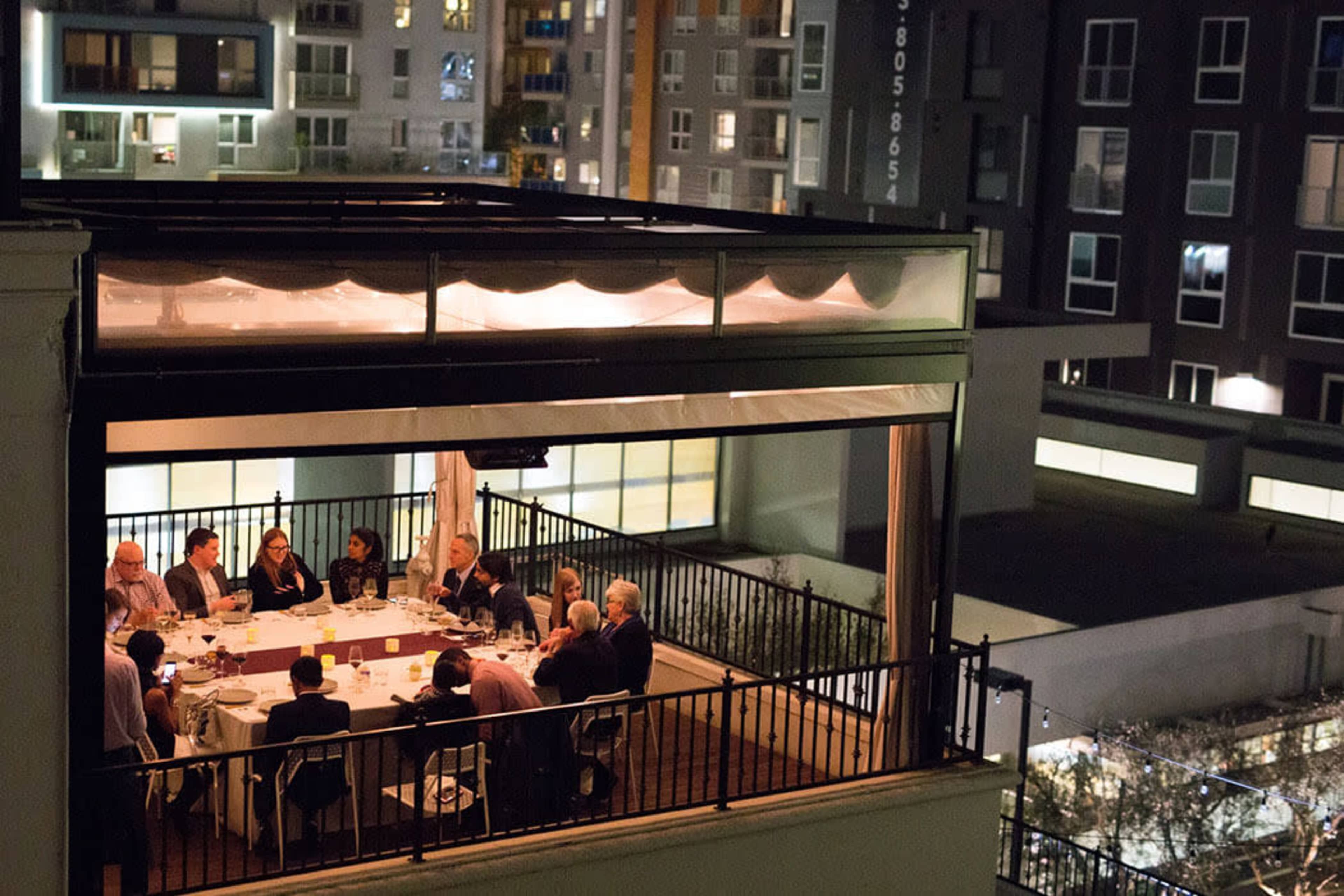 A group of people is dining together on a rooftop terrace, surrounded by city buildings at night.
