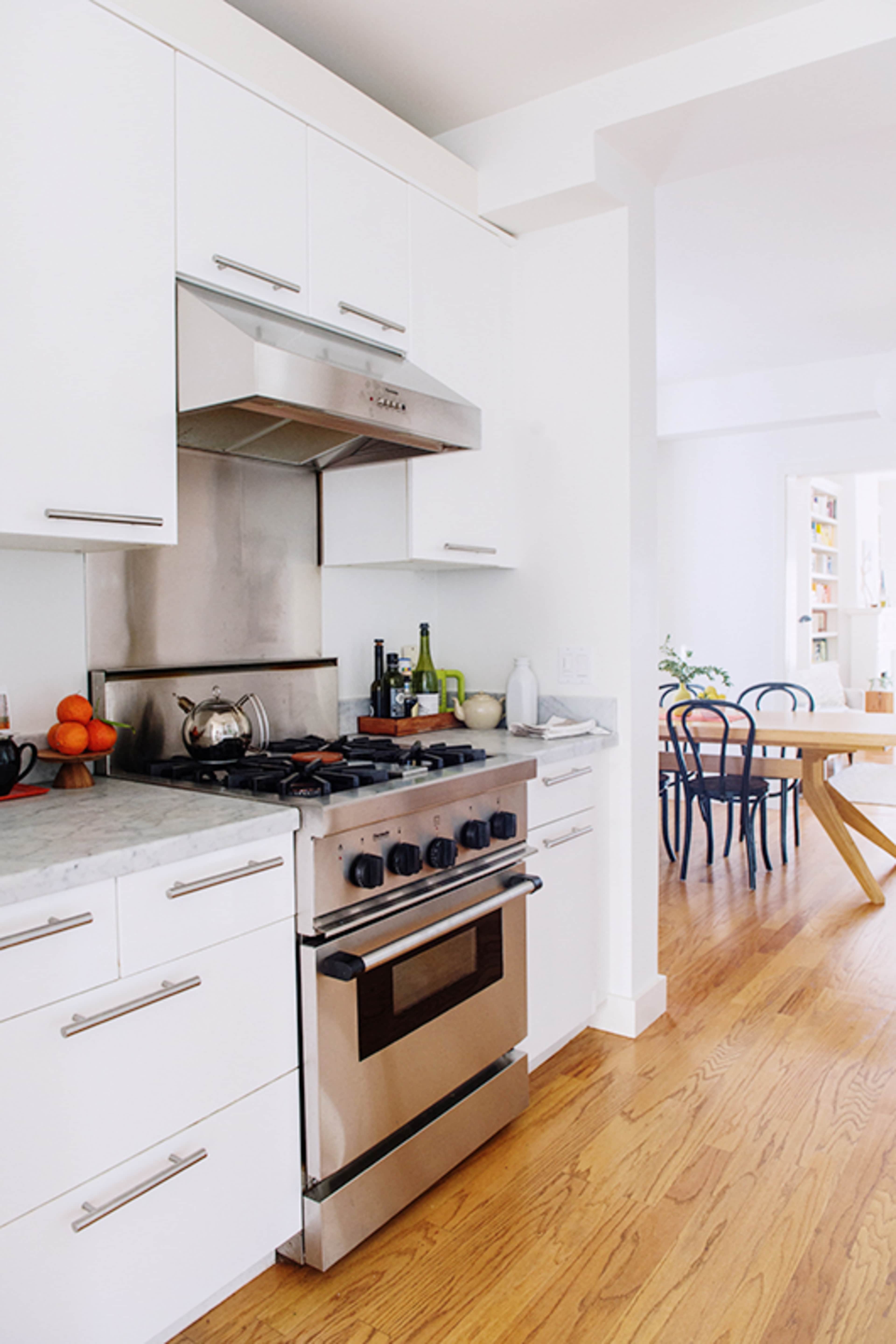 A modern kitchen with white cabinets, a stainless steel stove, and wooden floors, adjacent to a dining area with a wooden table and chairs.