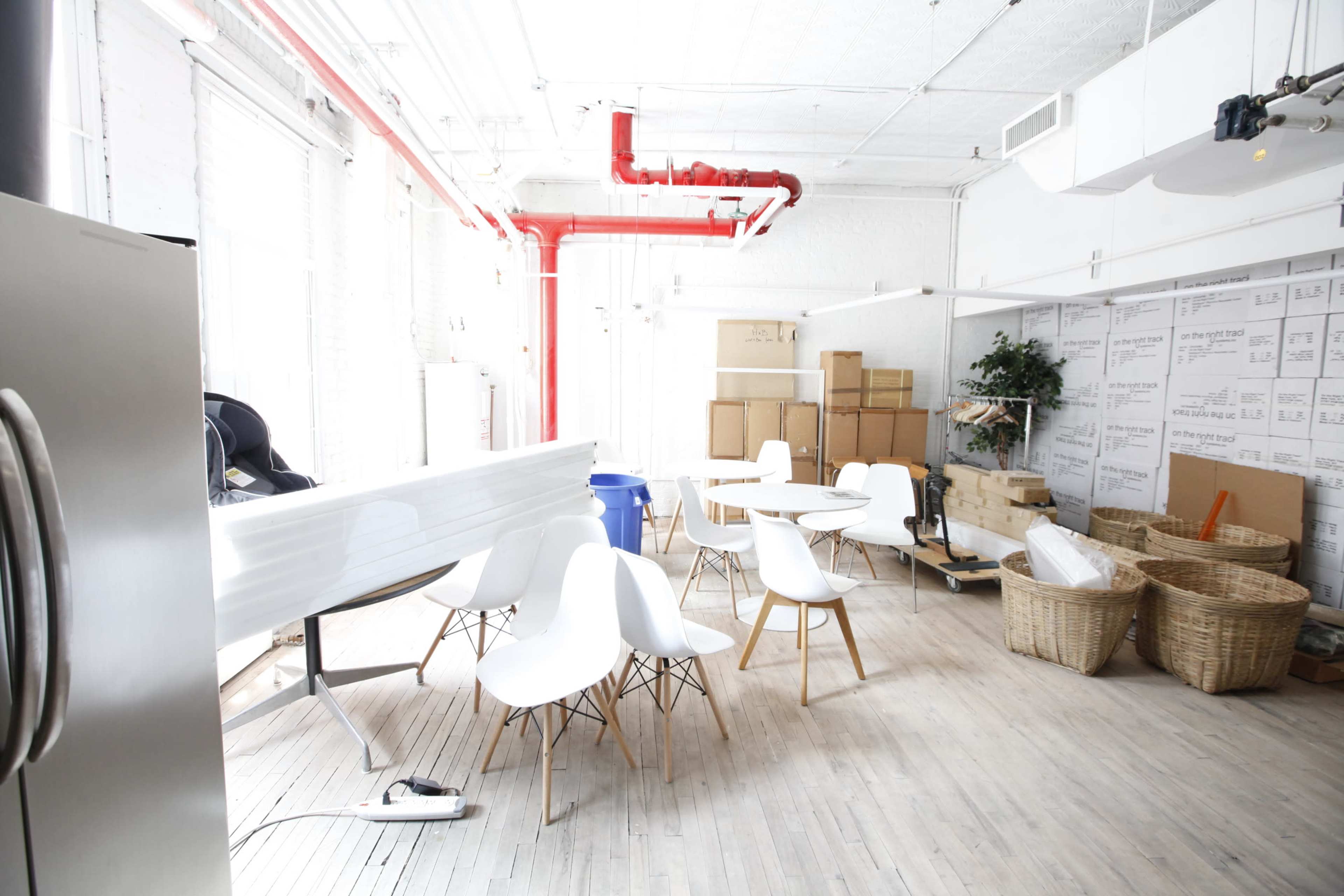 A bright, organized workspace featuring several white chairs around wooden tables, cardboard boxes stacked in the corner, and a few plants.