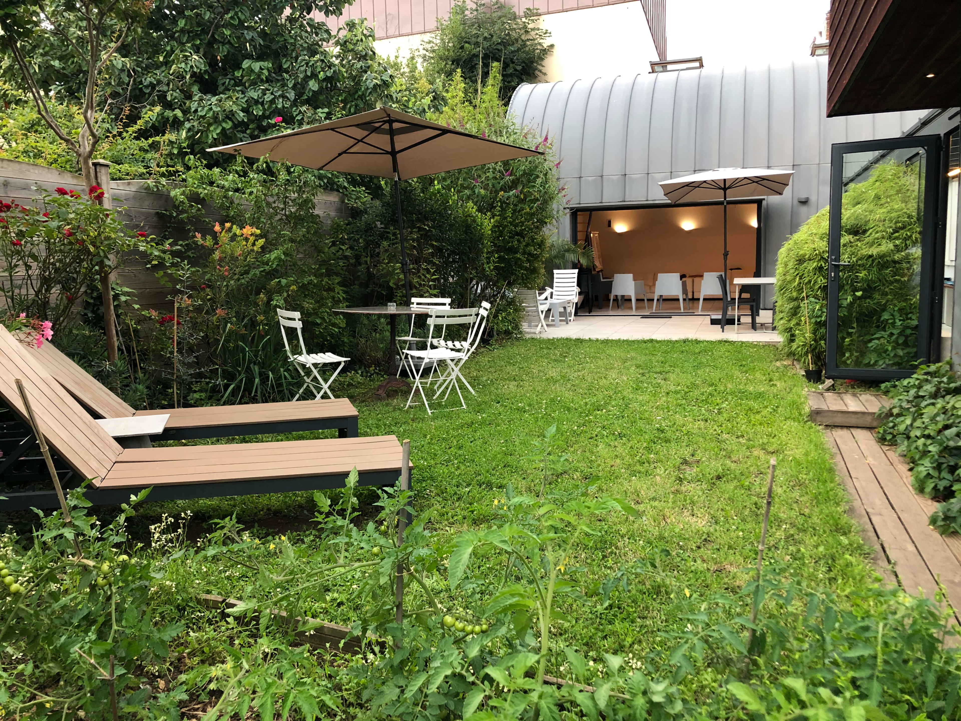 A well-maintained garden features green grass, two lounge chairs, a dining table with chairs, and two umbrellas, adjacent to a modern building.