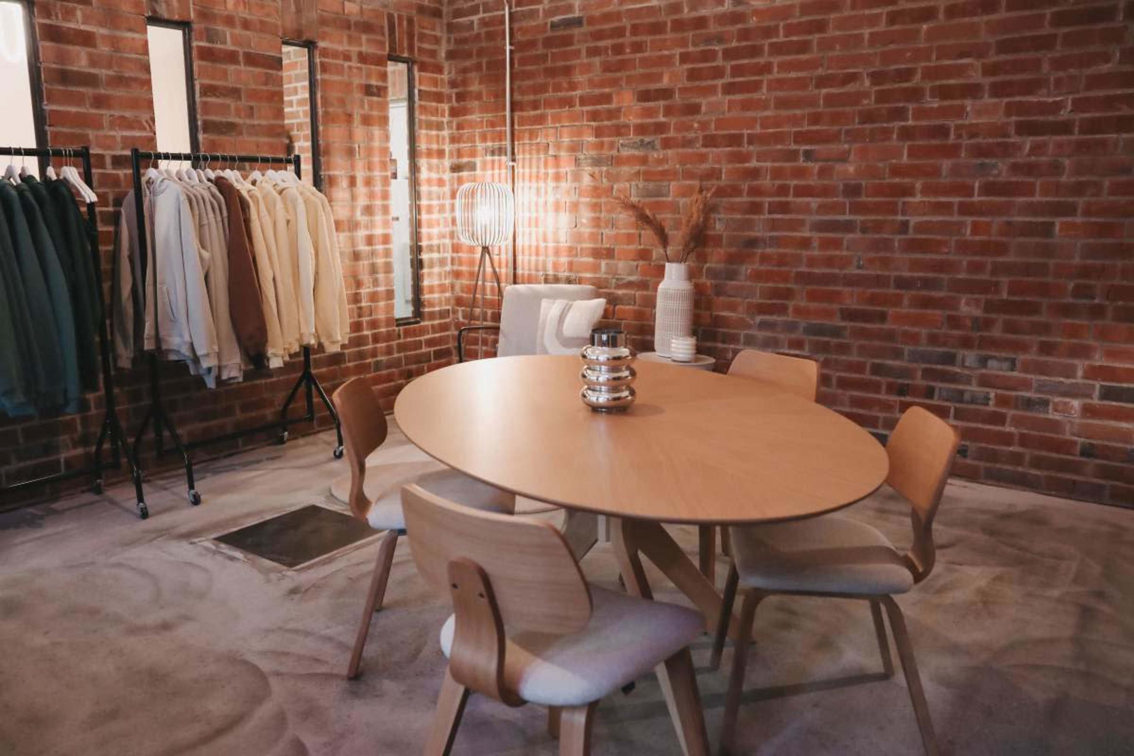 The image shows a circular wooden table surrounded by four wooden chairs in a room with exposed brick walls and clothing displayed on a rack.