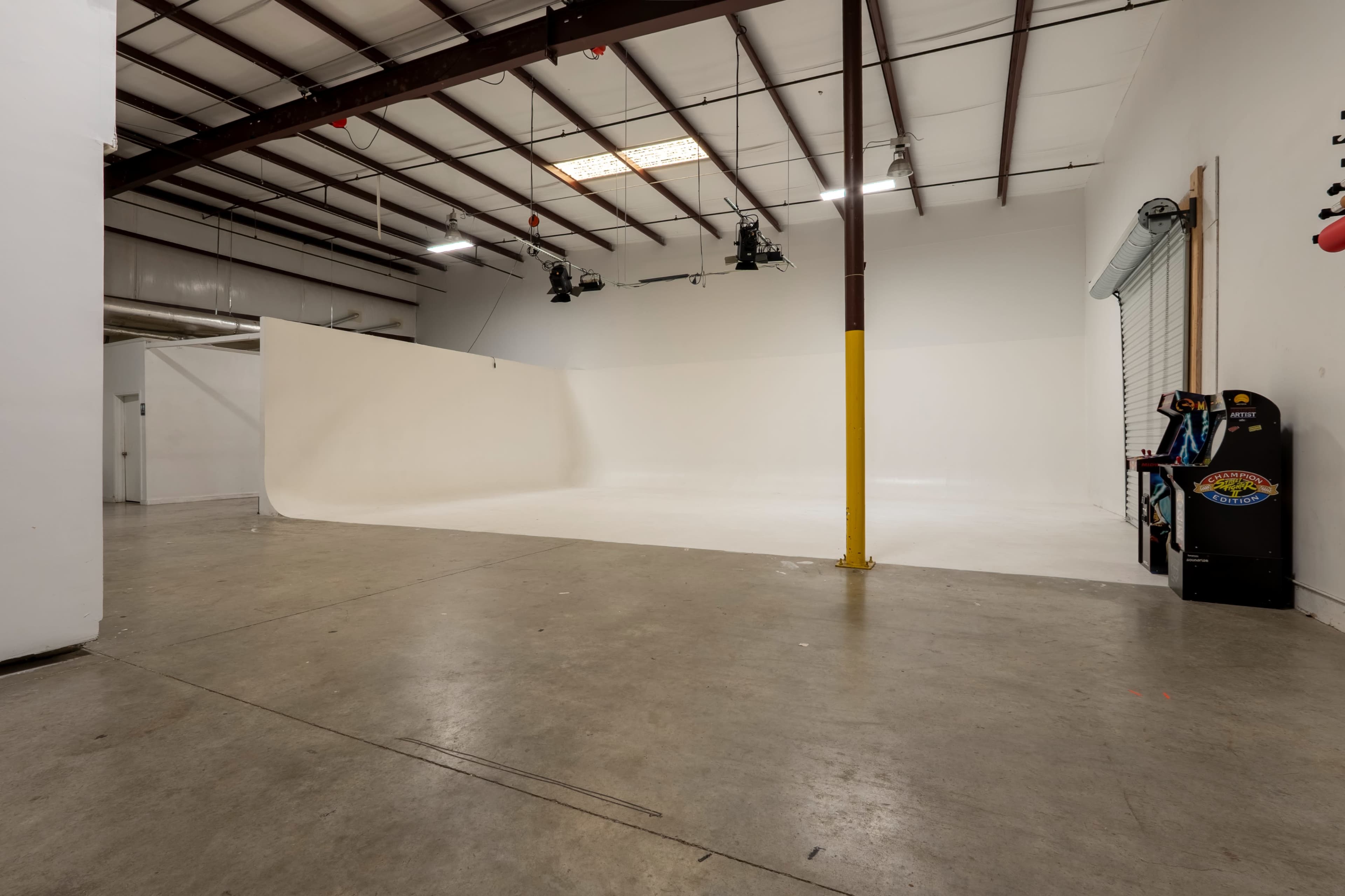The image shows a spacious indoor studio with a smooth white backdrop, concrete flooring, and minimal props, including an arcade machine in the corner.
