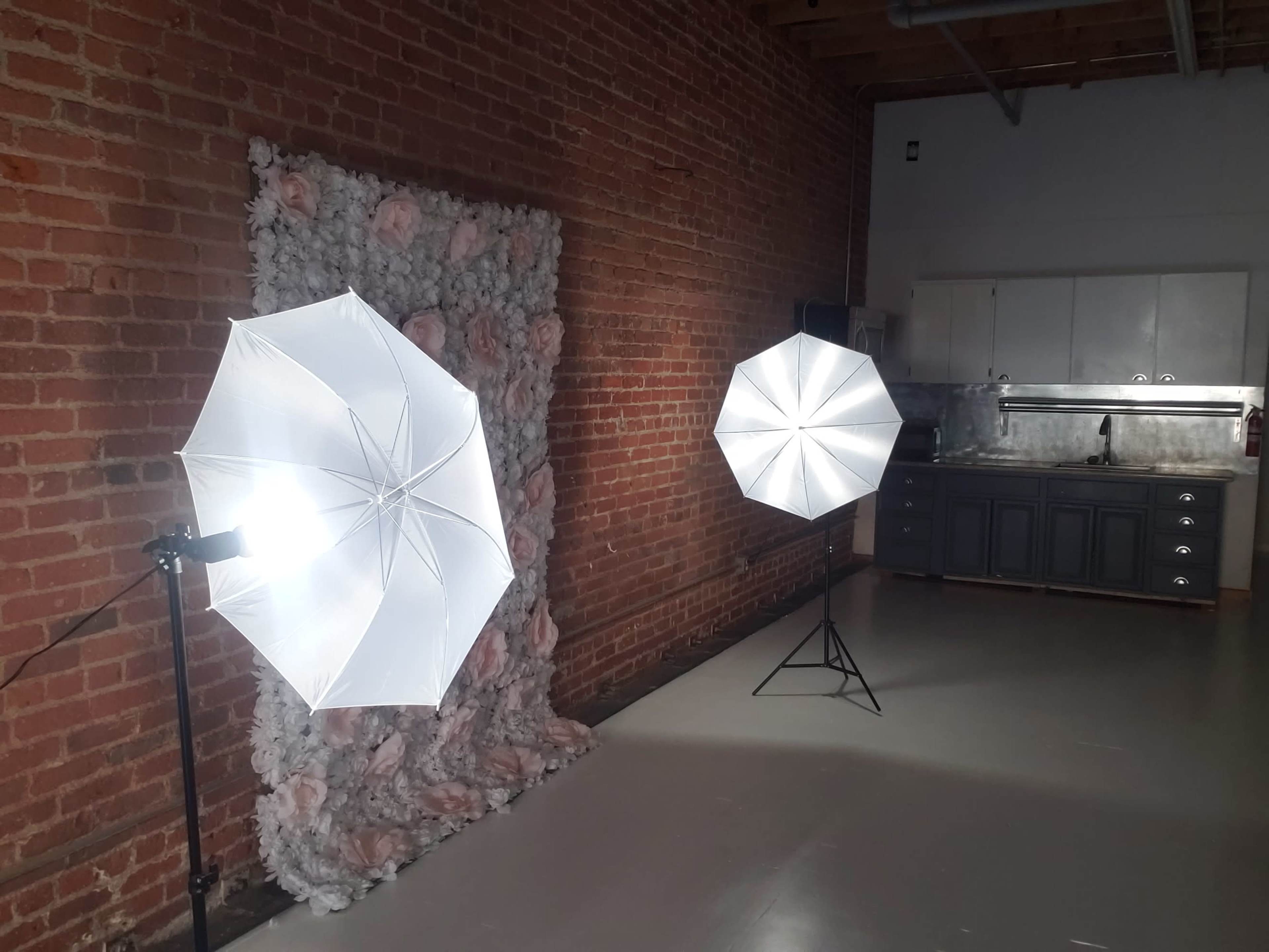 The image shows a photography setup with two softbox lights positioned on stands facing a textured backdrop in a room with exposed brick walls and a kitchenette in the background.