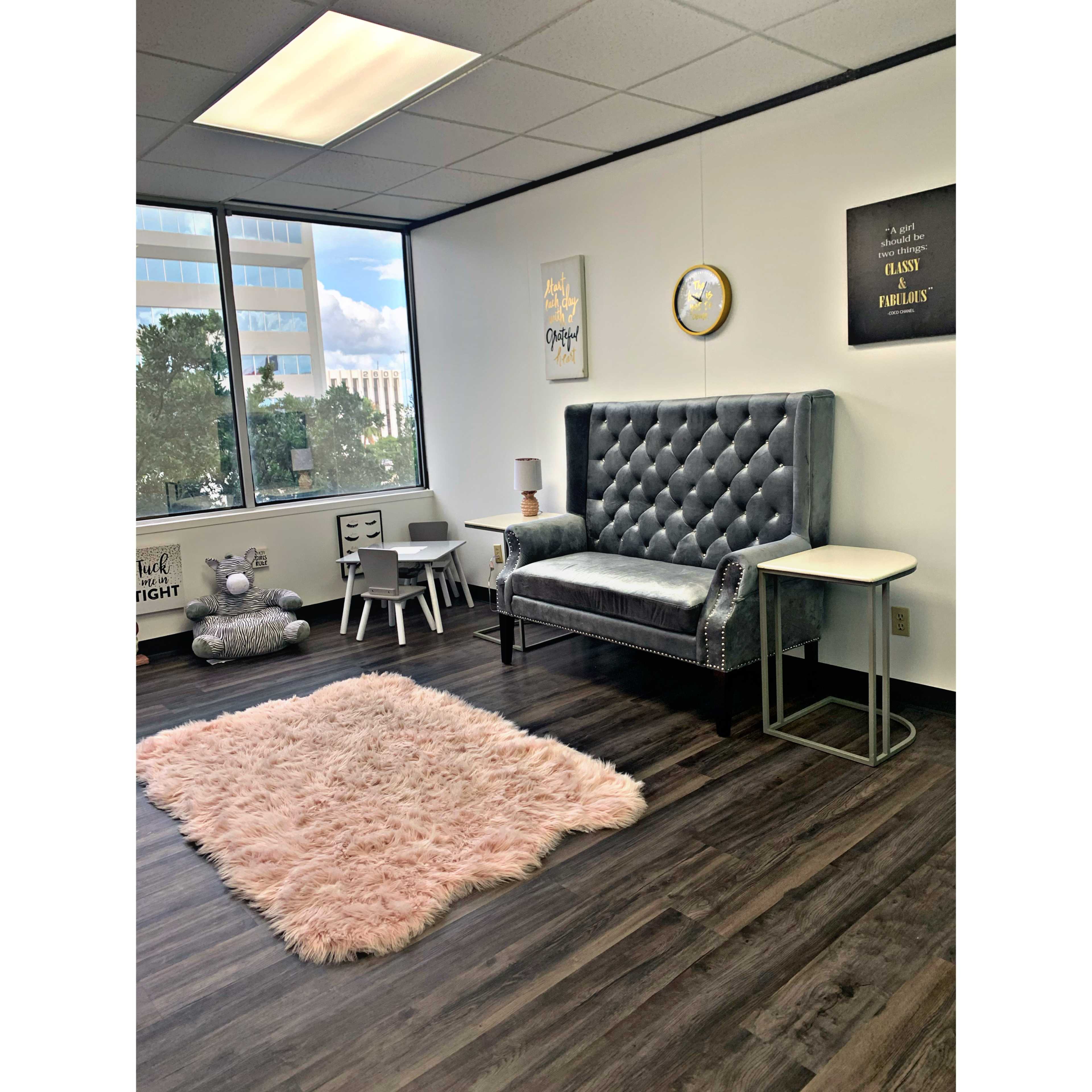 The image shows a modern office space featuring a plush gray armchair, a small white side table, a light pink rug, and a few minimalist furniture pieces beside a large window.