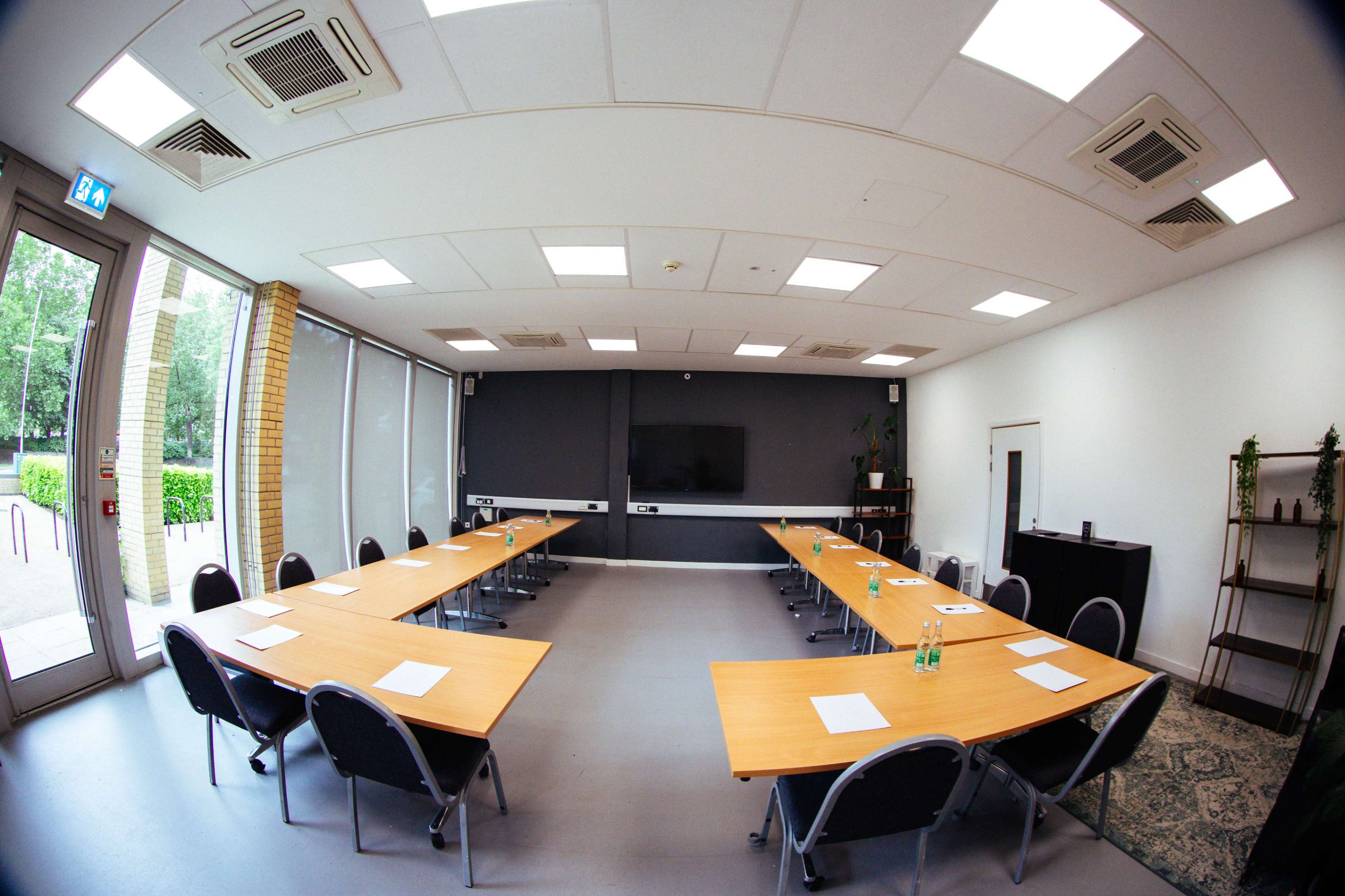 The image shows a conference room with two long tables arranged in a U-shape, surrounded by chairs, and equipped with a large screen on one wall.