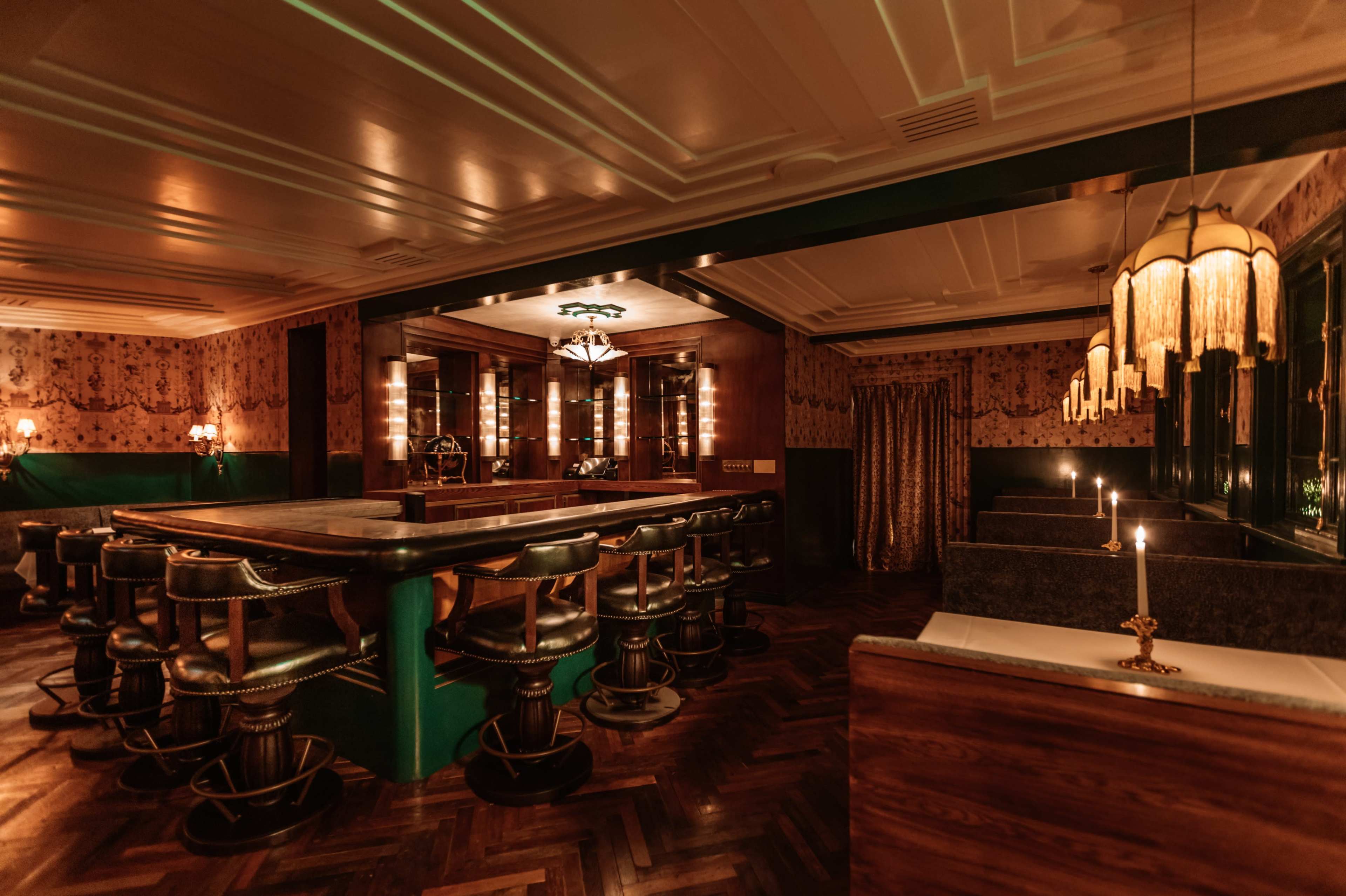 The image shows a dimly lit bar area with a wooden counter, high-backed stools, and elegant light fixtures, surrounded by patterned wallpaper and private booths.