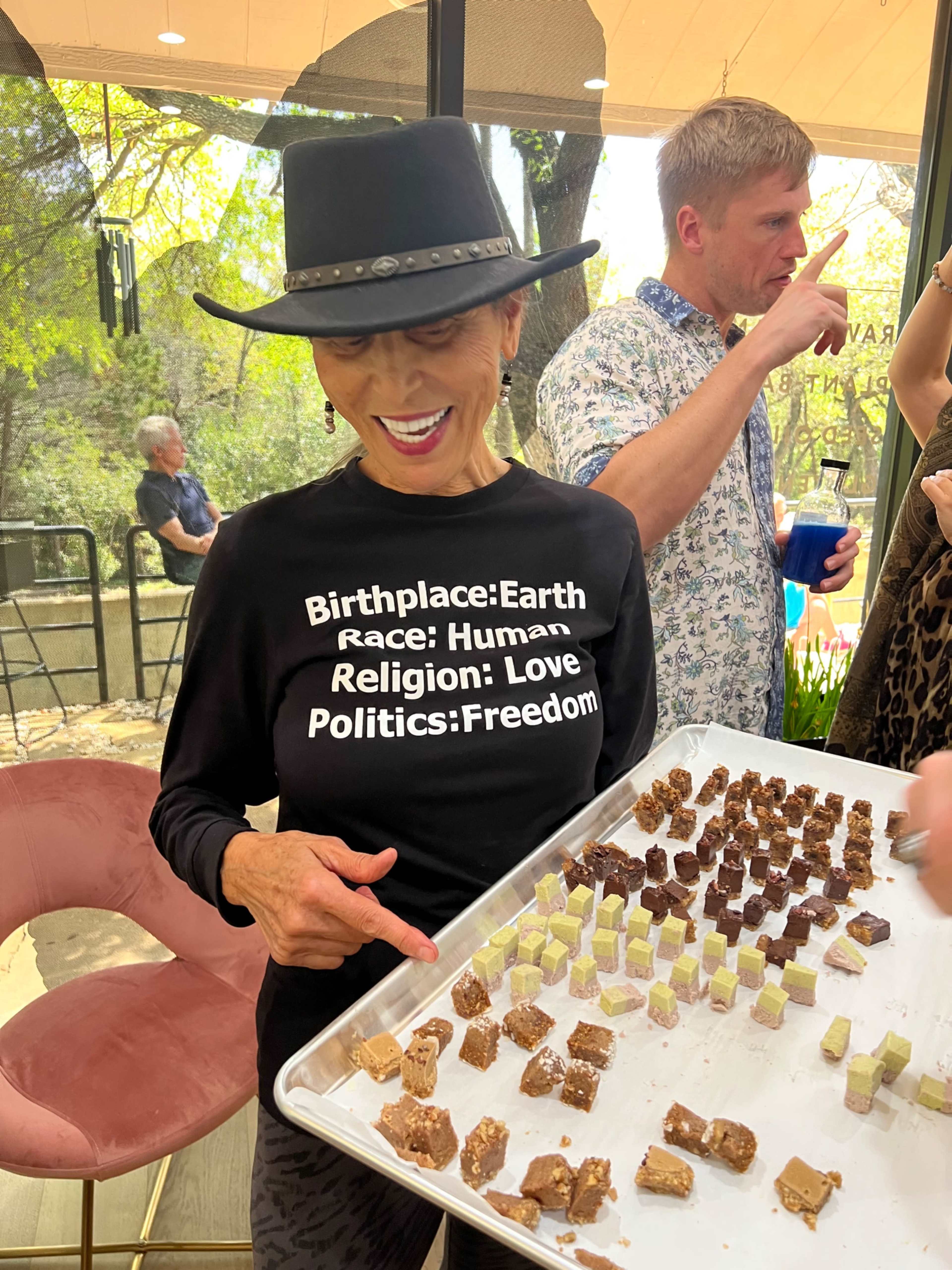 A woman wearing a black shirt with printed sayings holds a tray of assorted dessert squares while smiling at the camera.