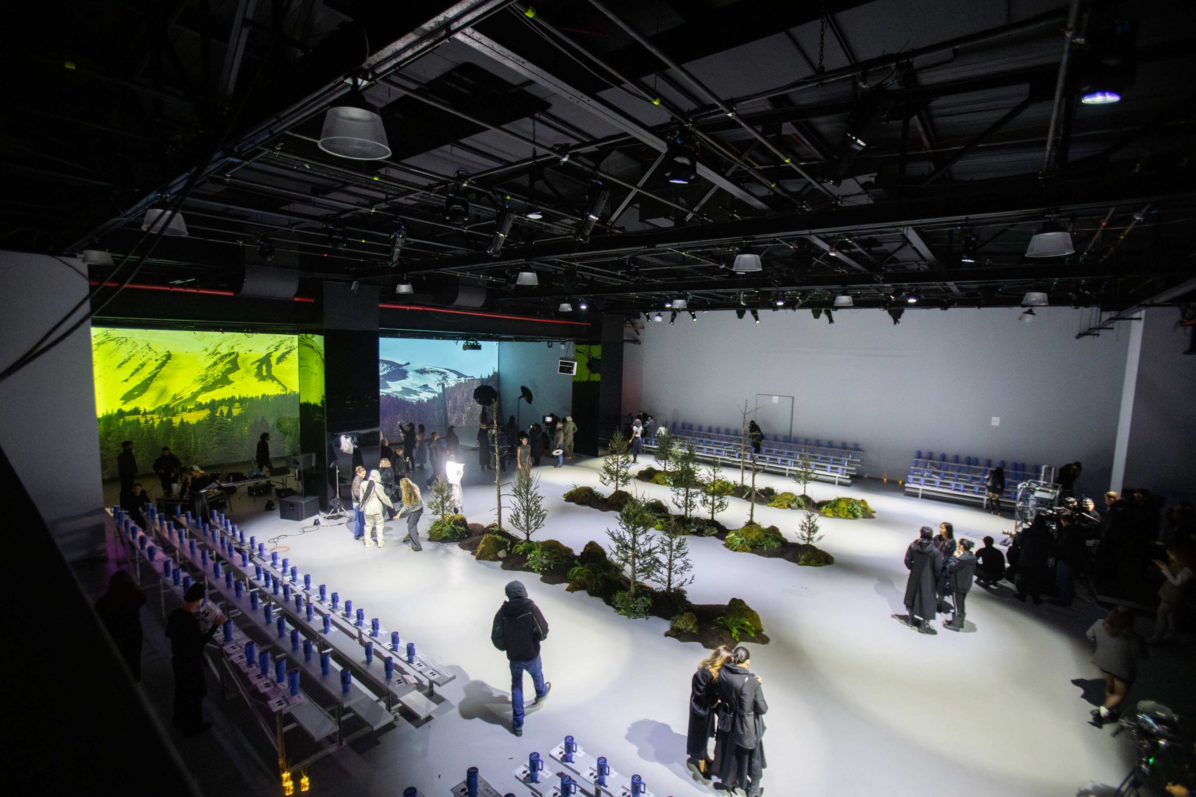 The image shows a fashion show venue with a minimalist design, featuring a white runway surrounded by neatly arranged greenery and seating for the audience.
