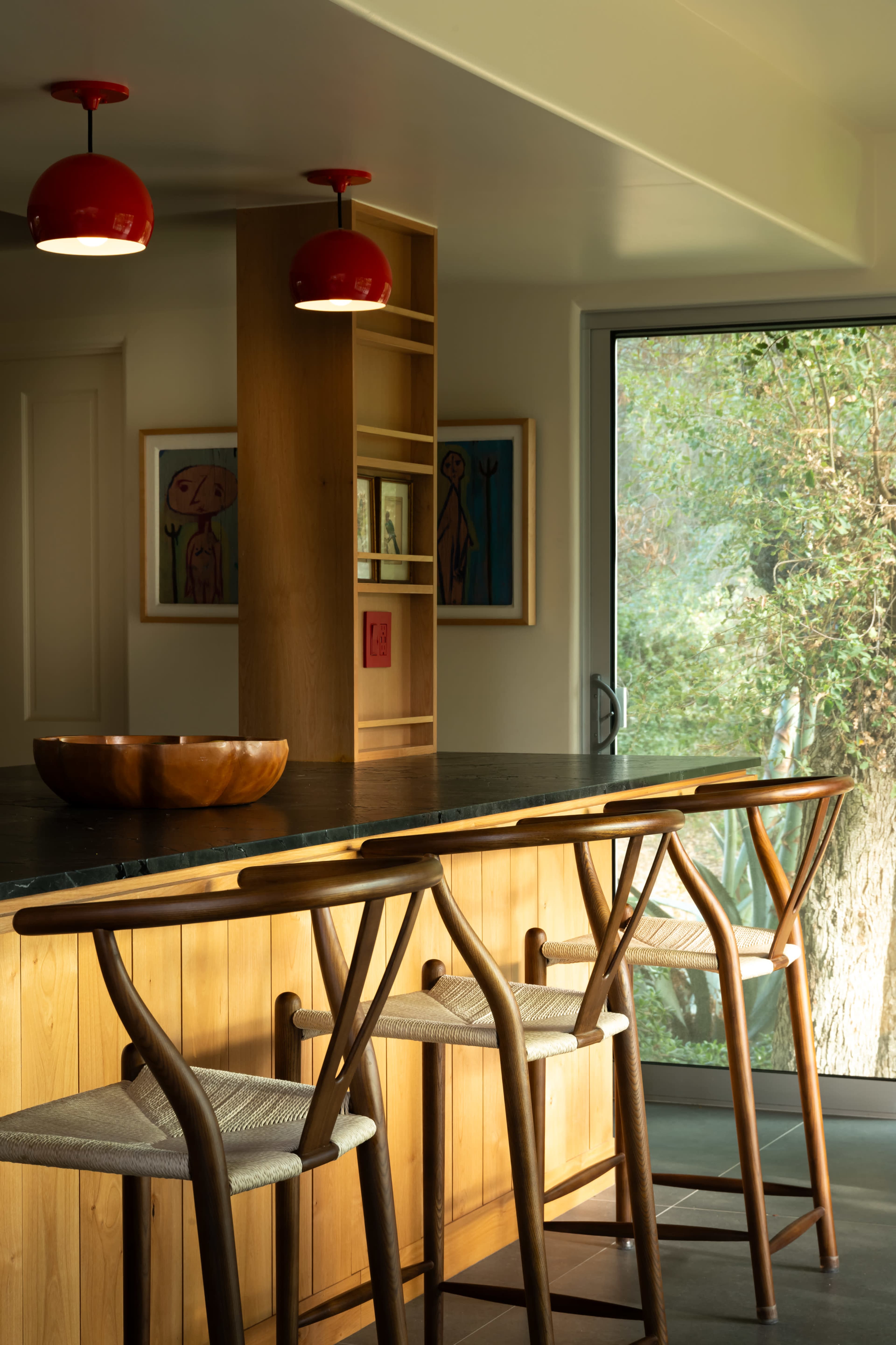 A modern kitchen features a wooden bar with three stools, red pendant lights overhead, and a large window displaying greenery outside.