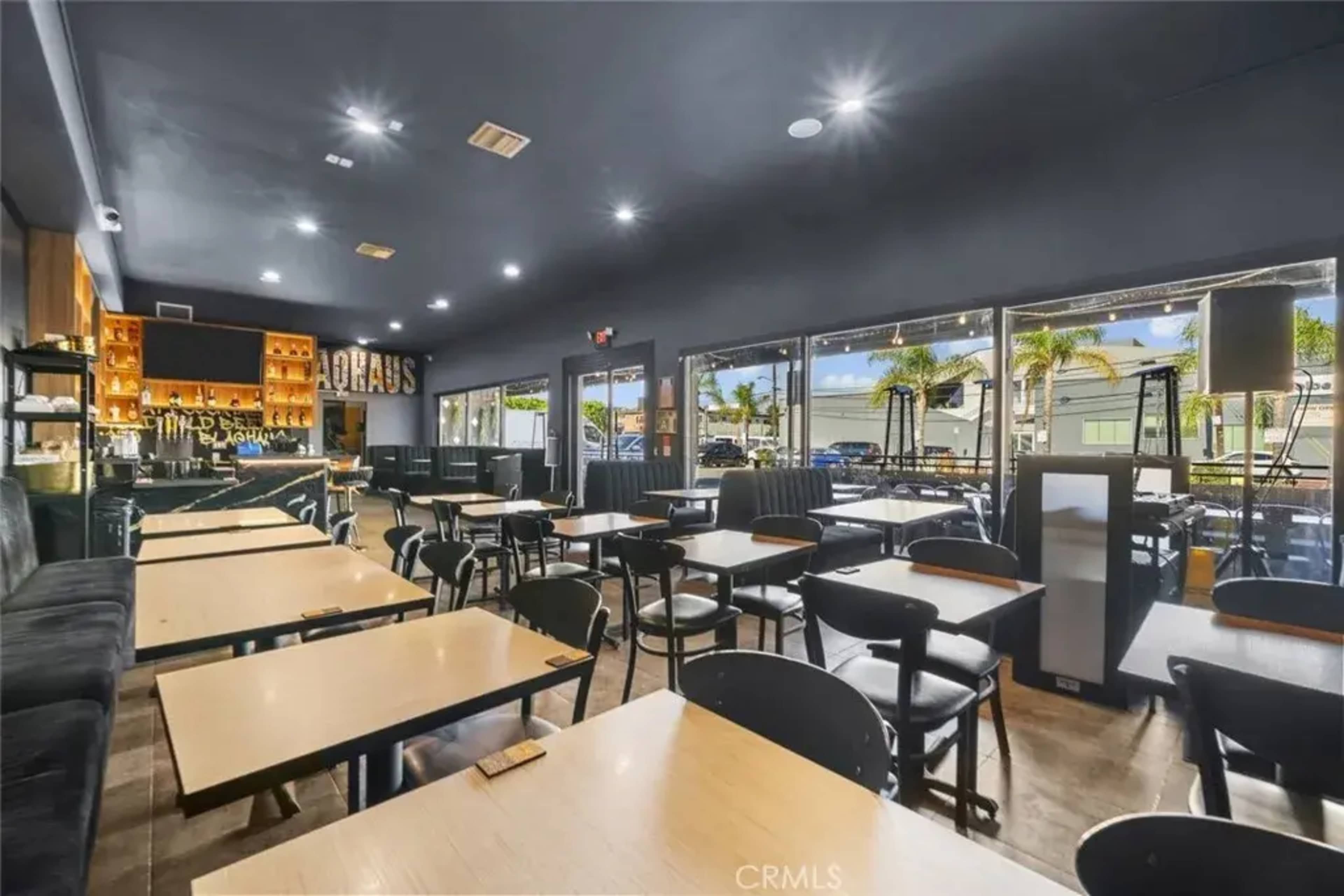 The image shows an interior of a modern restaurant with wooden tables, black chairs, and a bar area in the back.