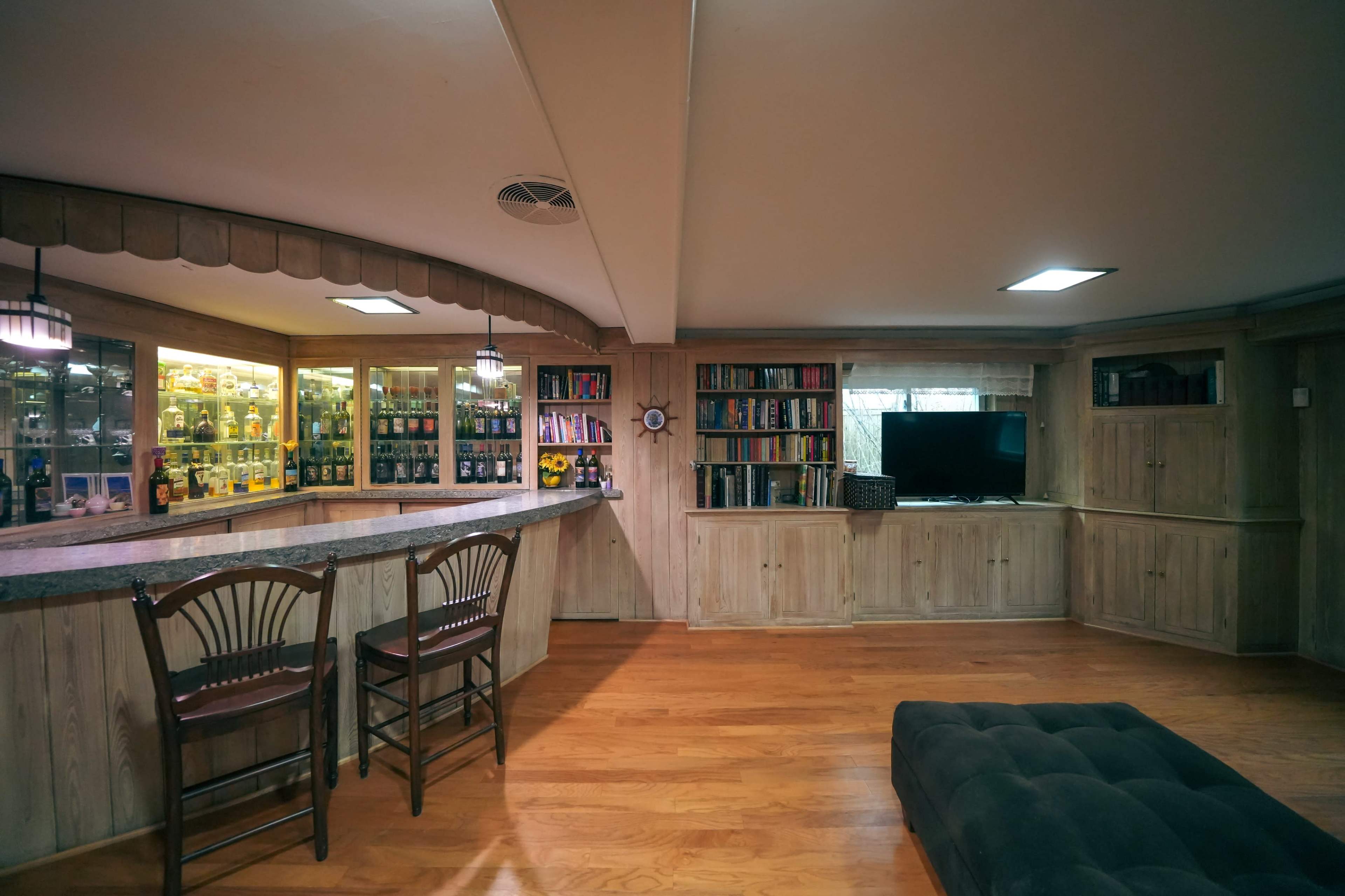A cozy basement with a wooden bar, shelves of books, and a television, all illuminated by overhead lights.