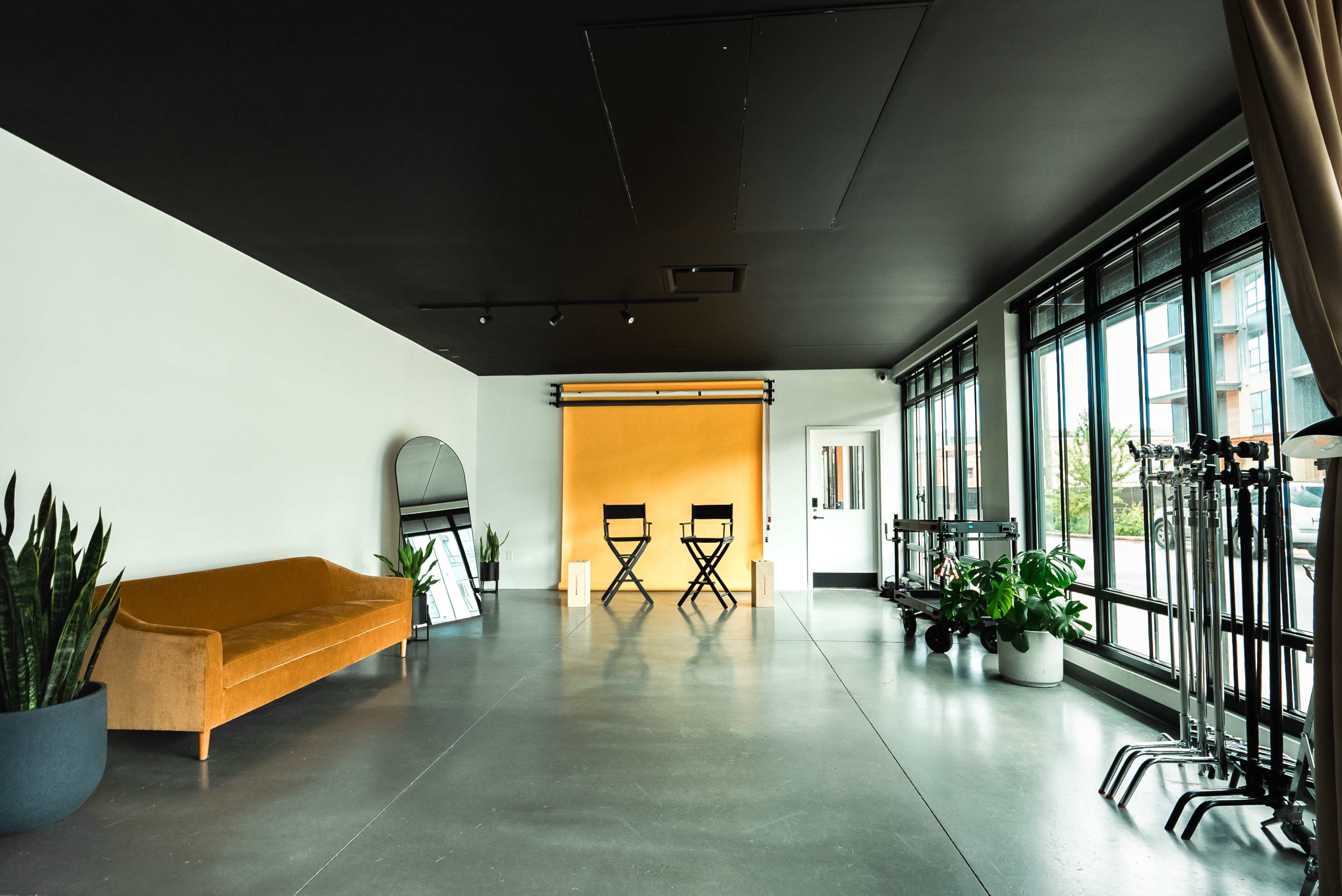 A spacious interior features a sofa, two chairs, a large mirror, and potted plants, with a backdrop and photography equipment against one wall.
