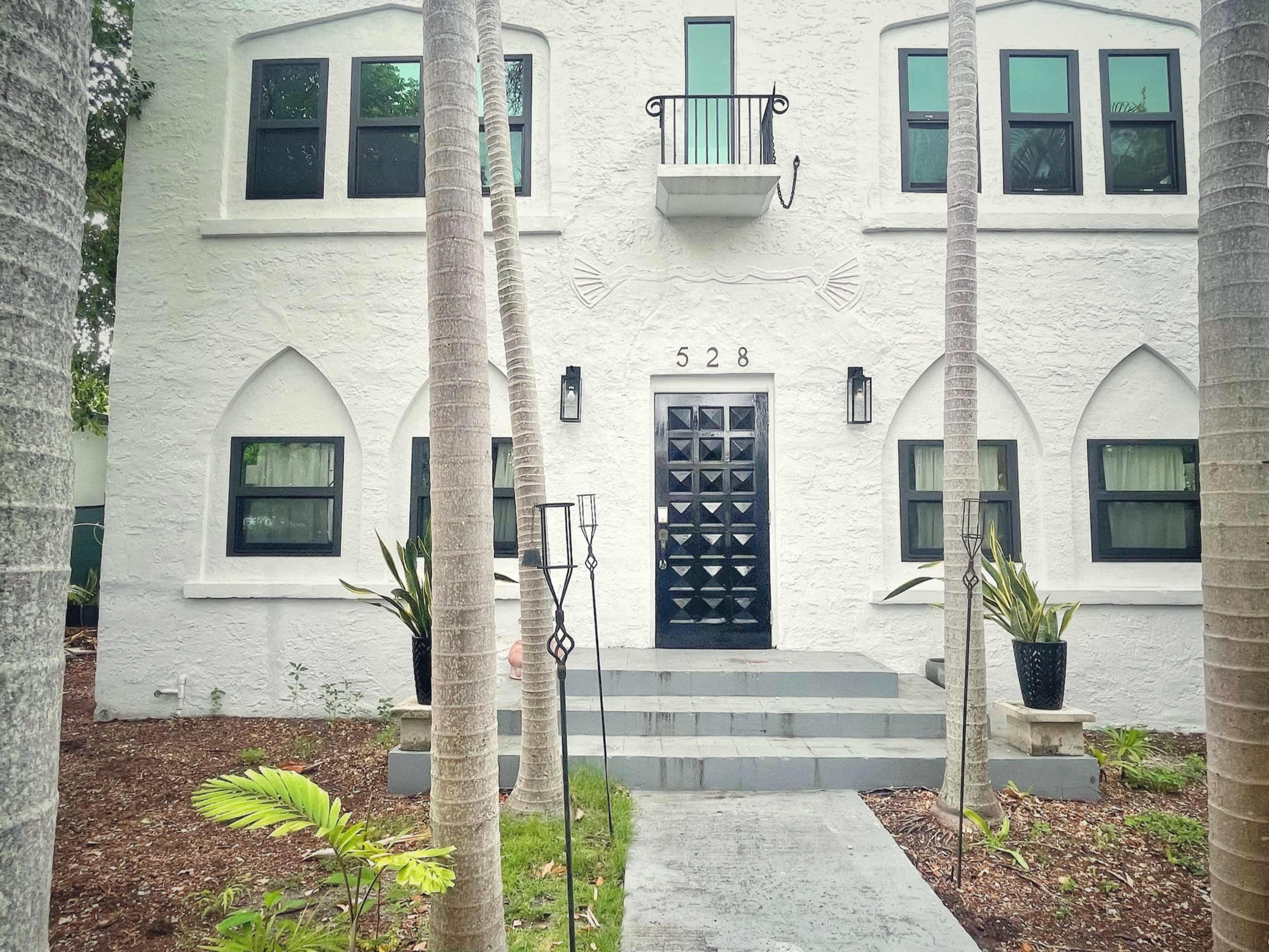 The image features a two-story white building with an ornate black door, flanked by palm trees and a pathway leading to the entrance.
