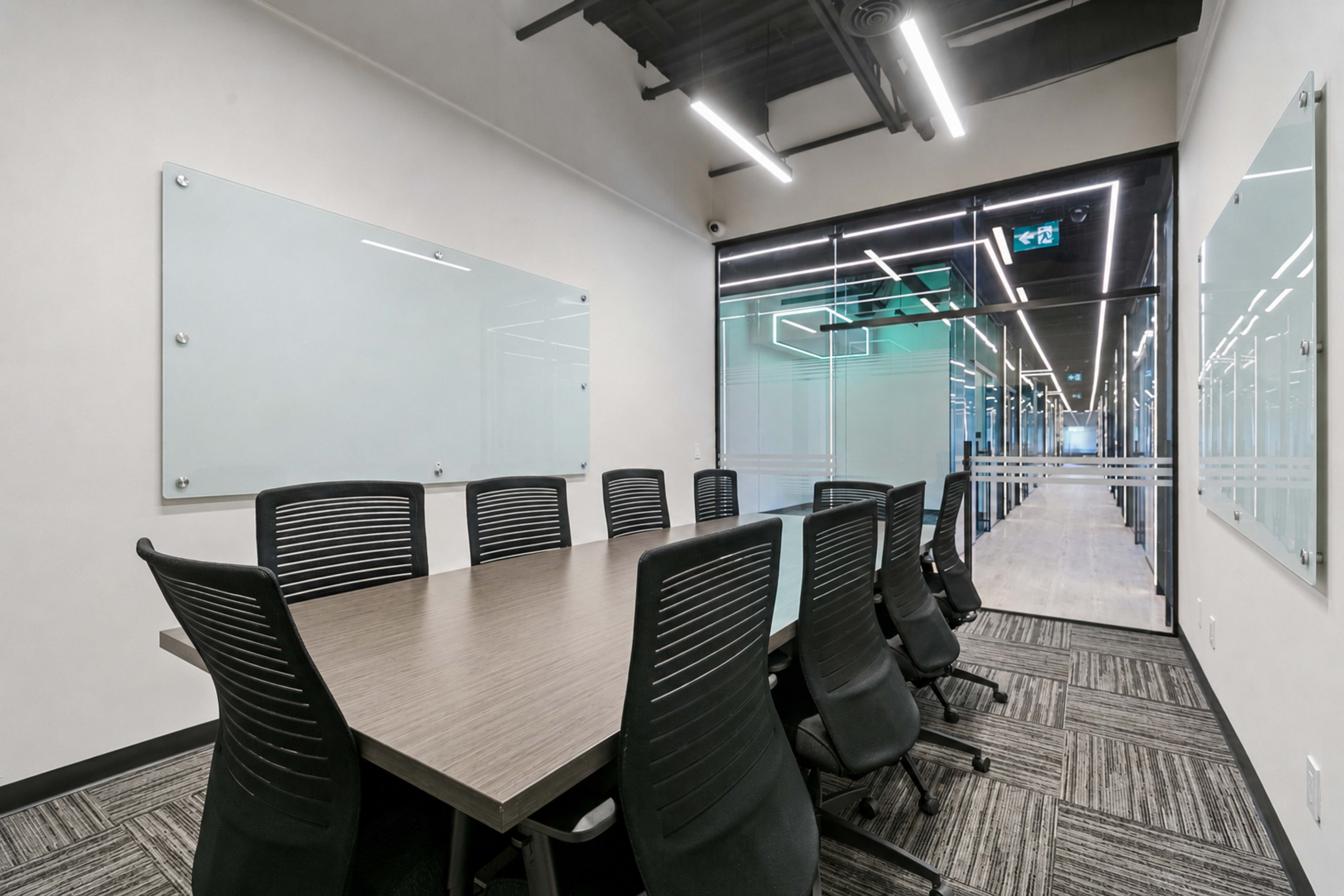 The image shows a modern conference room featuring a large wooden table surrounded by black ergonomic chairs, with glass panels on the walls.
