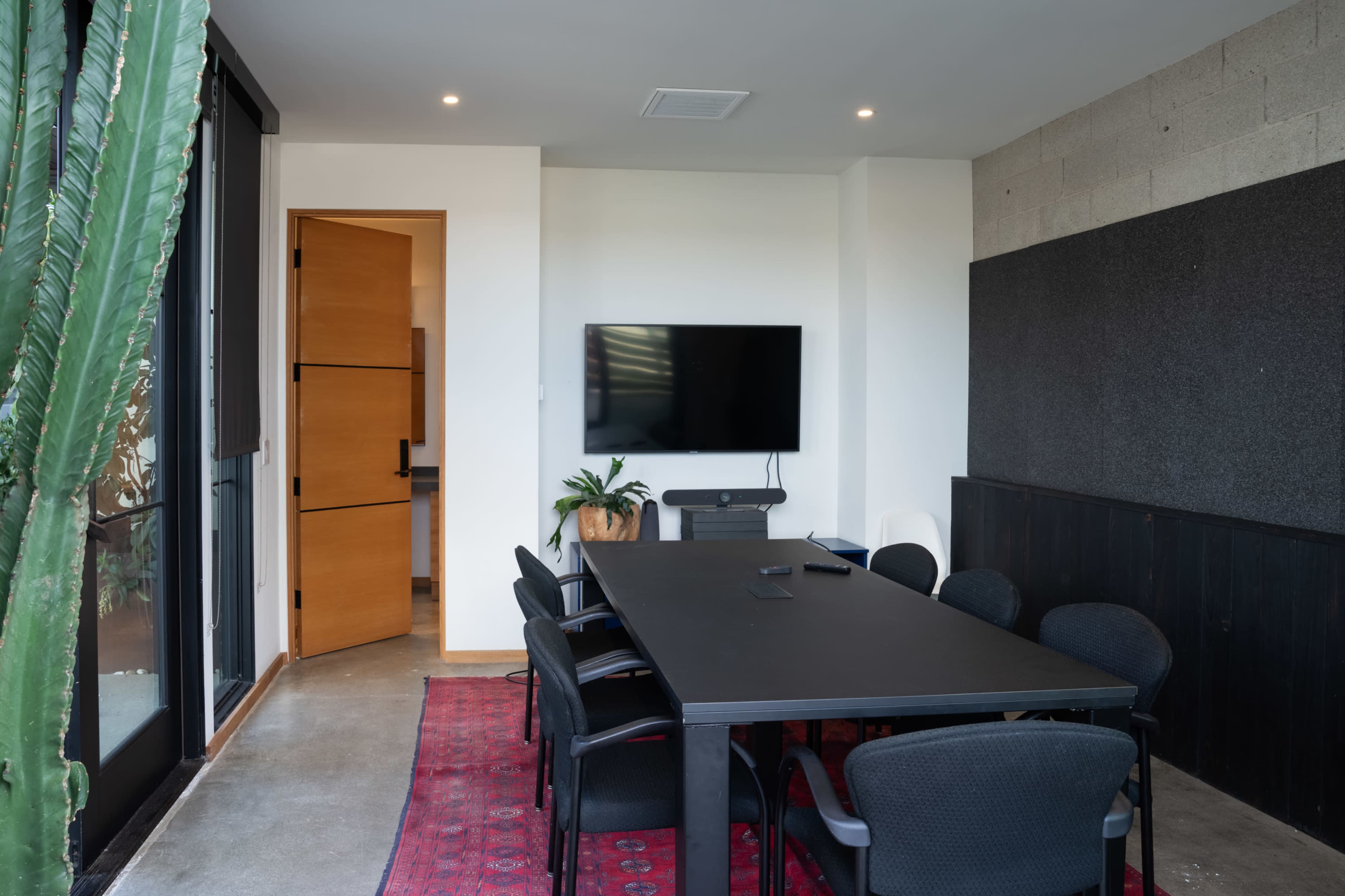 A modern conference room features a long black table surrounded by chairs, a television mounted on the wall, and a doorway leading to another room, with a large cactus in the corner.