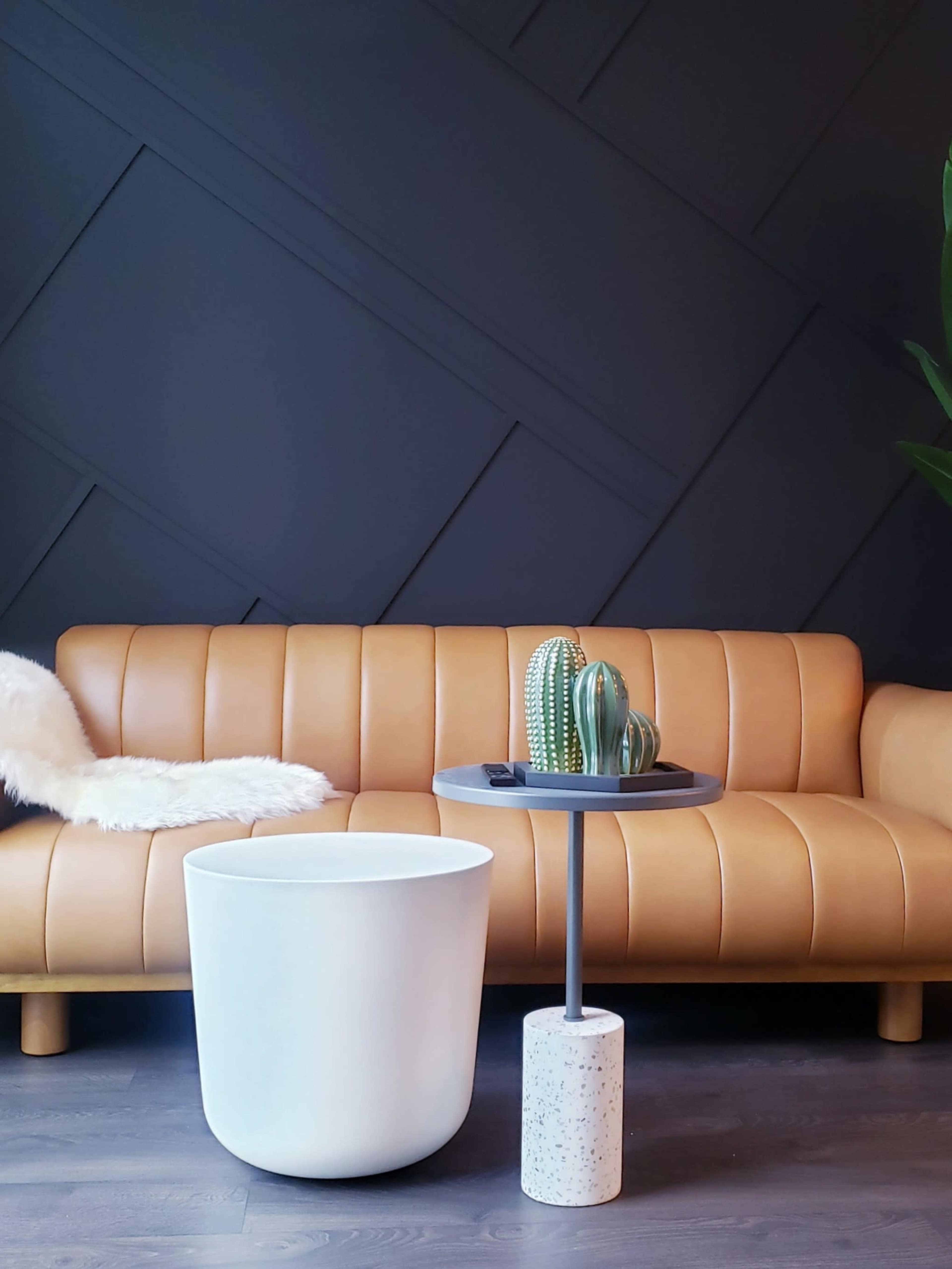 A tan leather sofa with a white side table and a small cactus arrangement on top, set against a dark geometric wall.