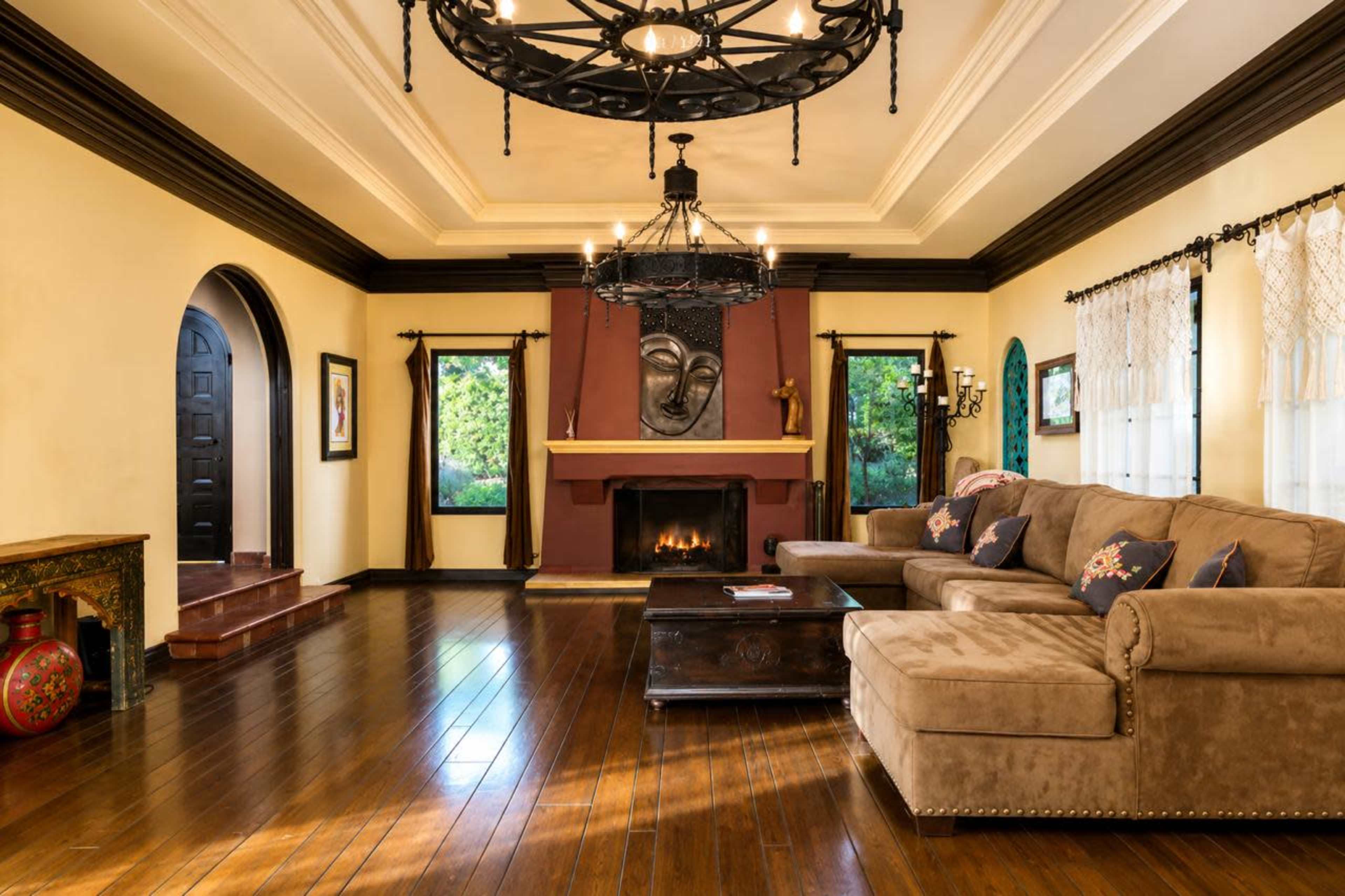 The image features a spacious living room with a tan sectional sofa, a wooden coffee table, a fireplace, and large windows that allow natural light to enter the space.
