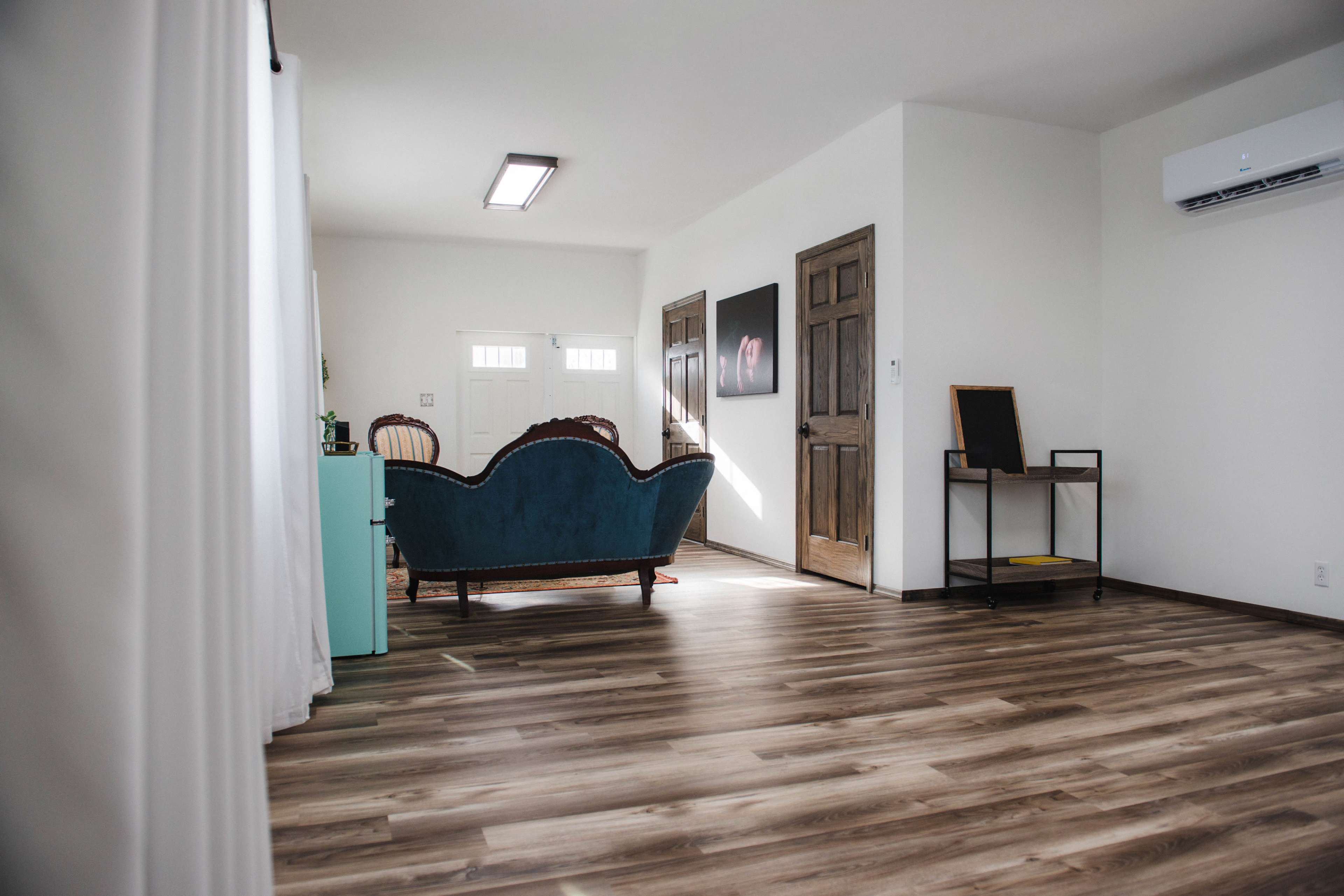A blue couch is positioned in a minimalist room with wooden floors and doors leading to an entryway.