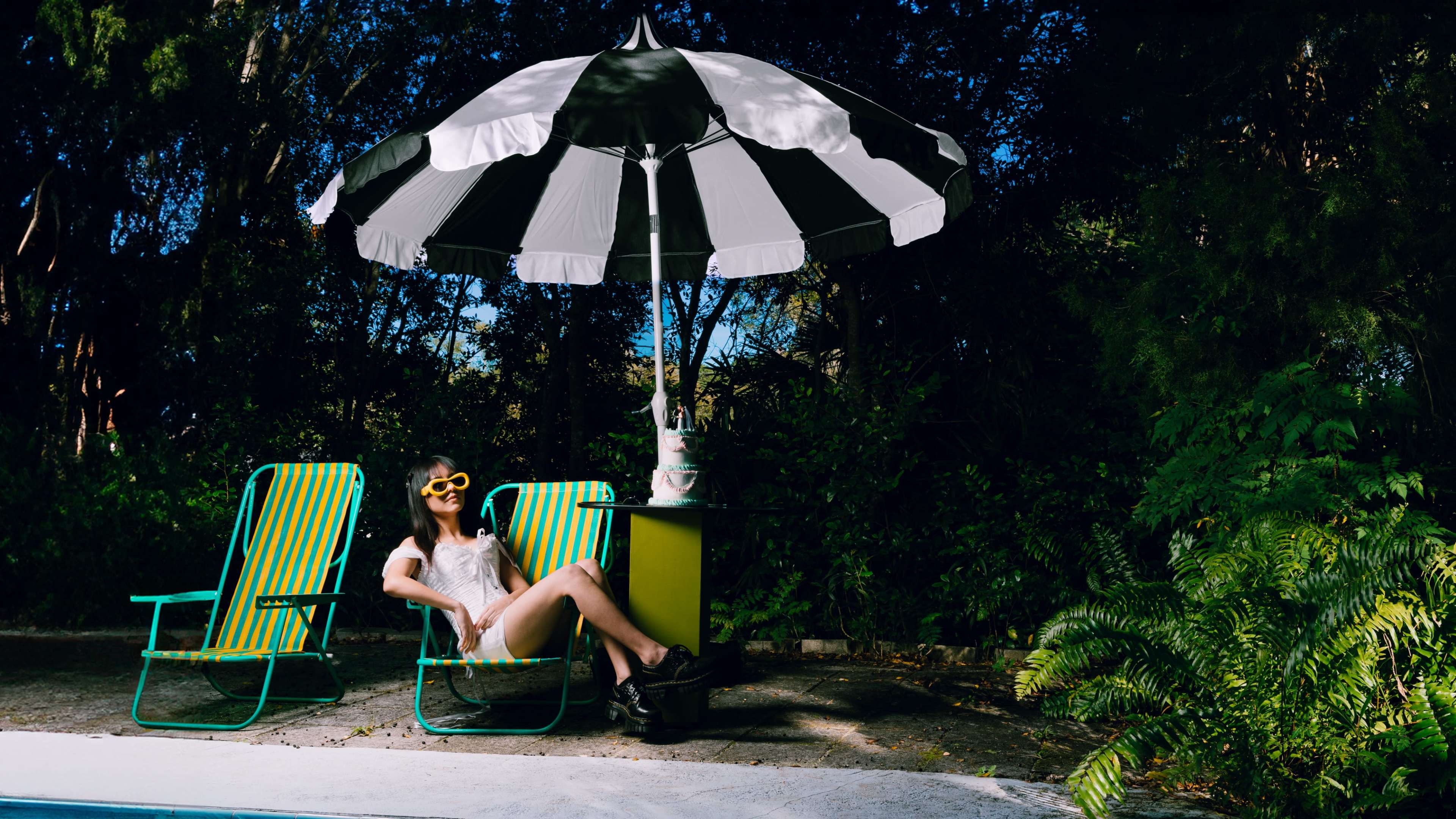A person relaxes in a striped deck chair under a large umbrella by a pool, surrounded by greenery.