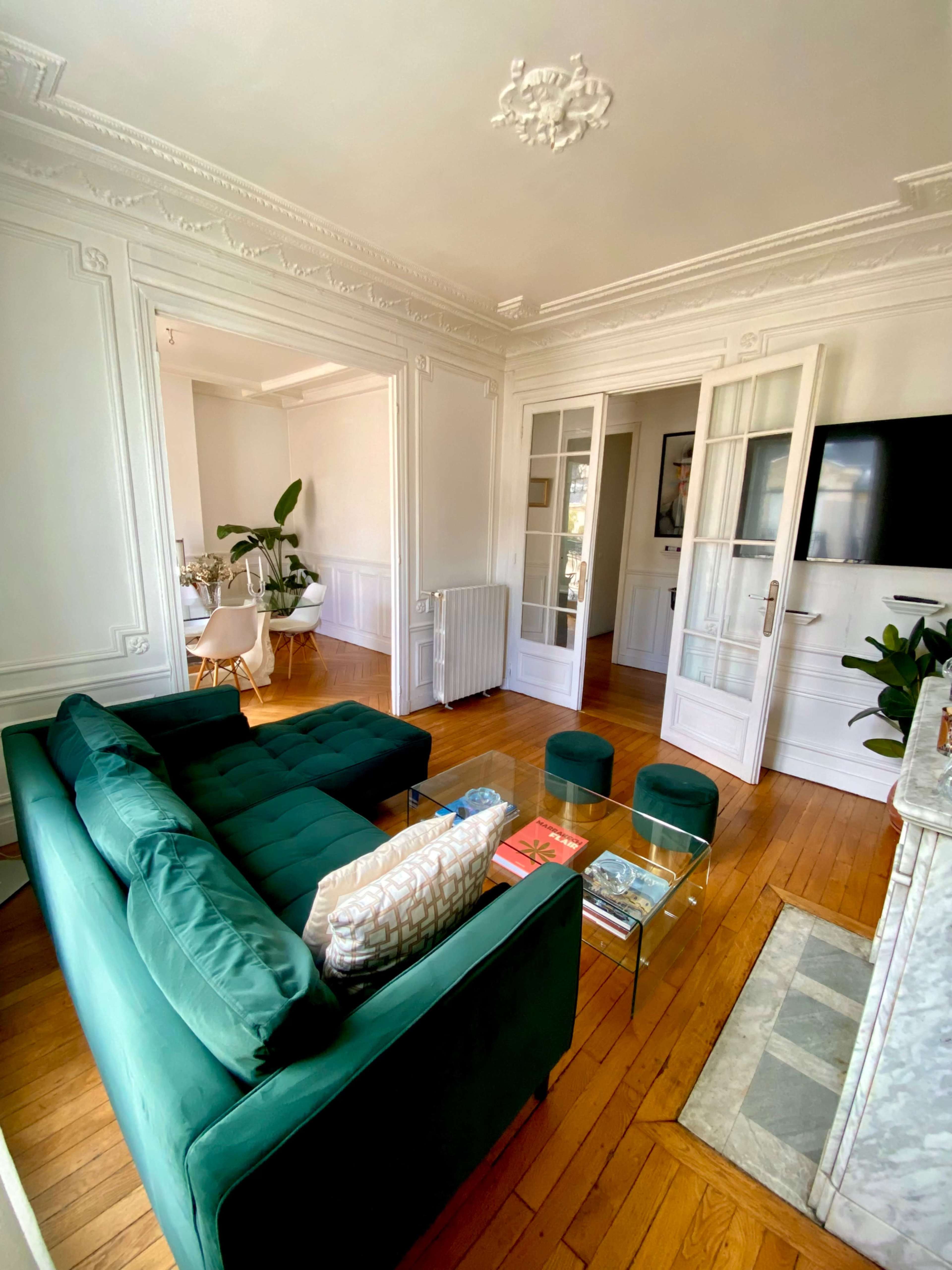 The living room features a green velvet sofa, glass coffee table, and hardwood floors, with a dining area visible through a set of French doors.