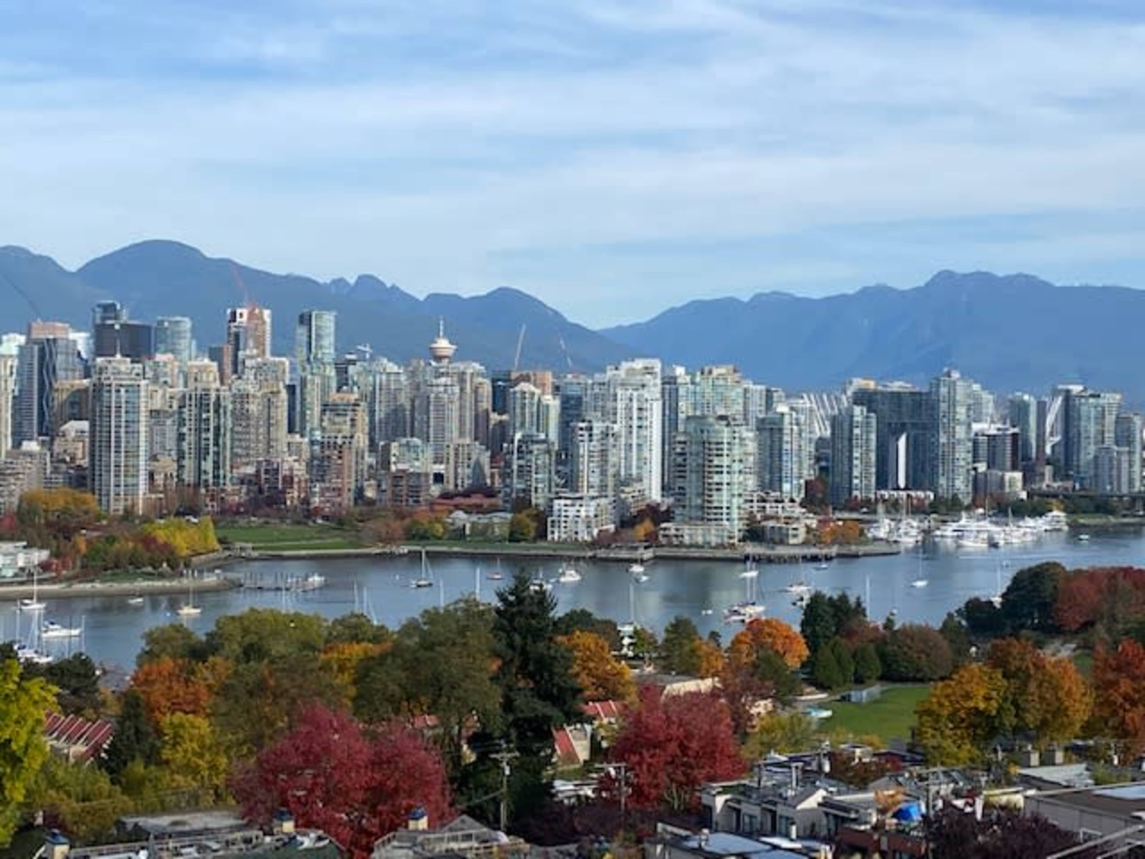 The image showcases a wide view of Vancouver's skyline, featuring tall buildings, a river, and distant mountains with autumn-colored trees in the foreground.