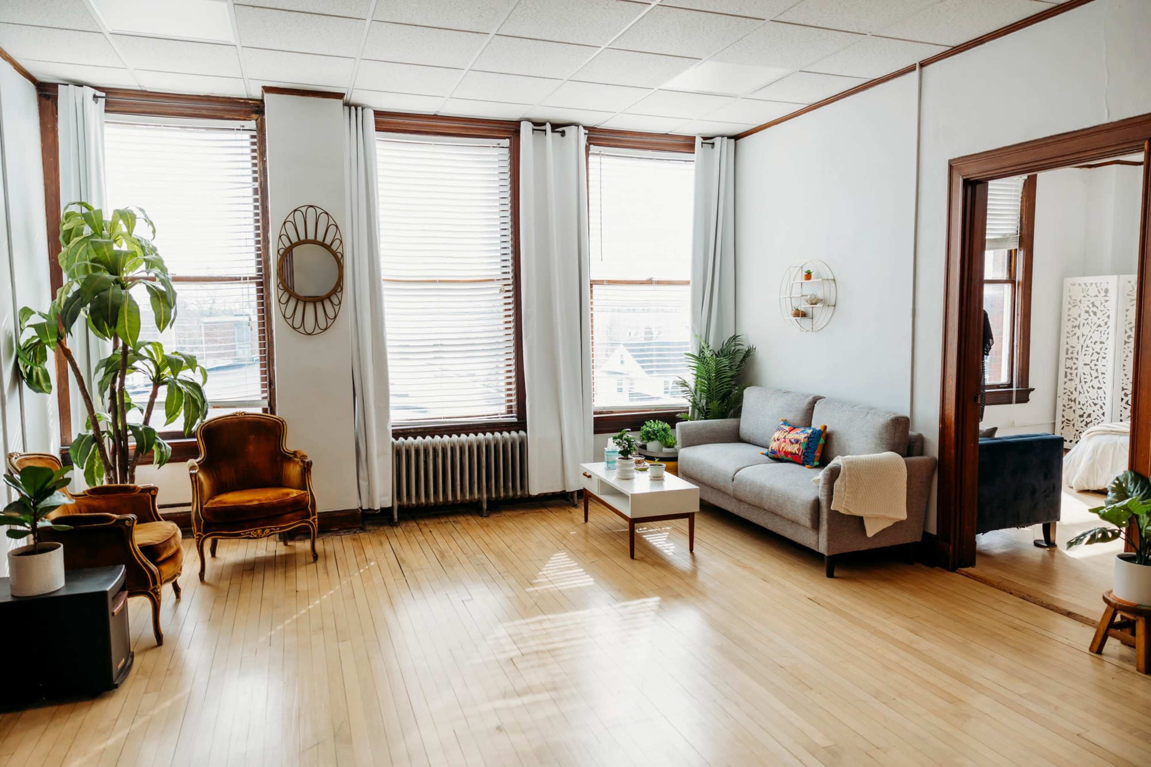 A bright living room features large windows, a gray sofa, wooden furniture, and indoor plants.