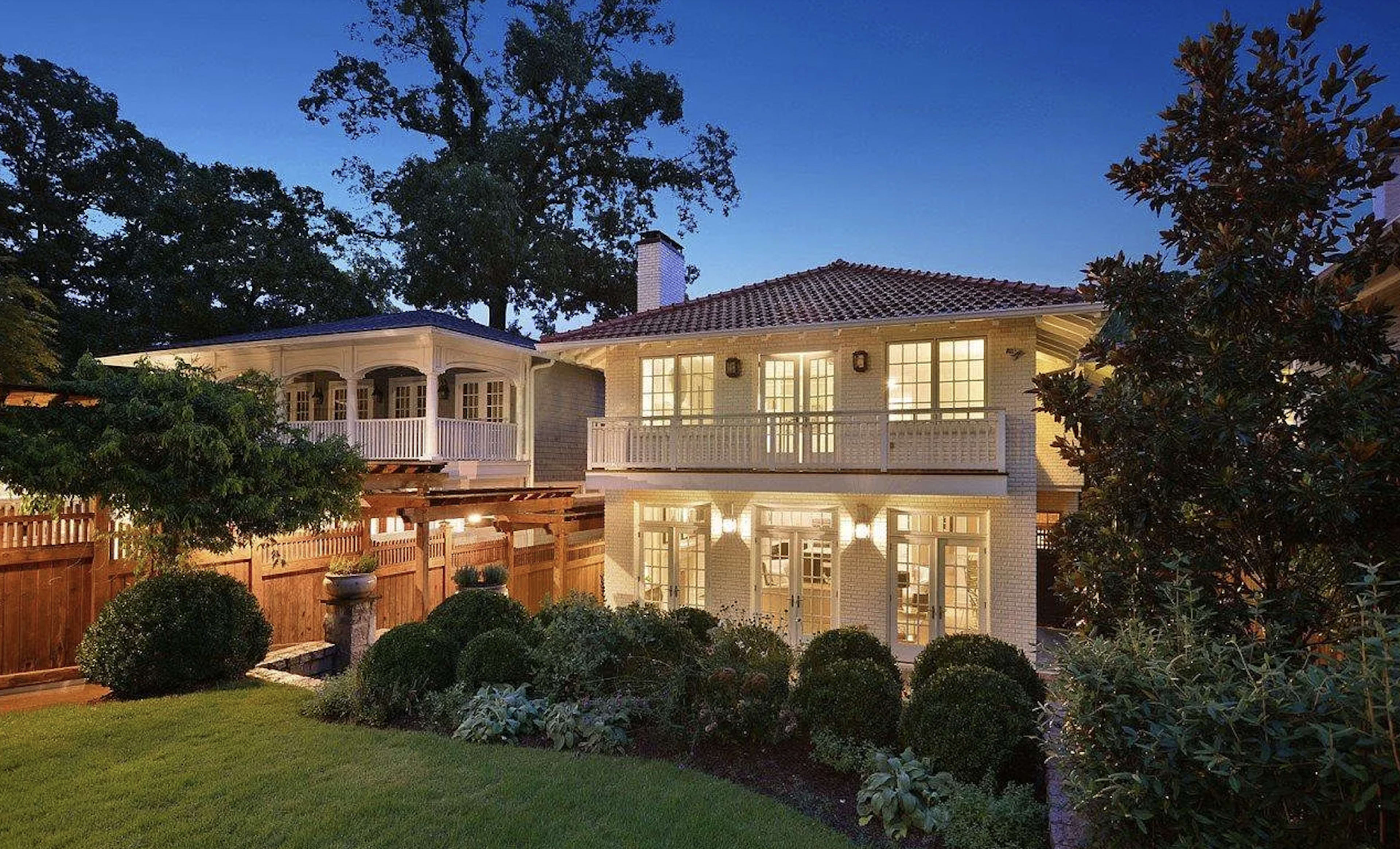 The image shows a well-lit two-story house with a tiled roof, surrounded by greenery and a wooden fence at dusk.