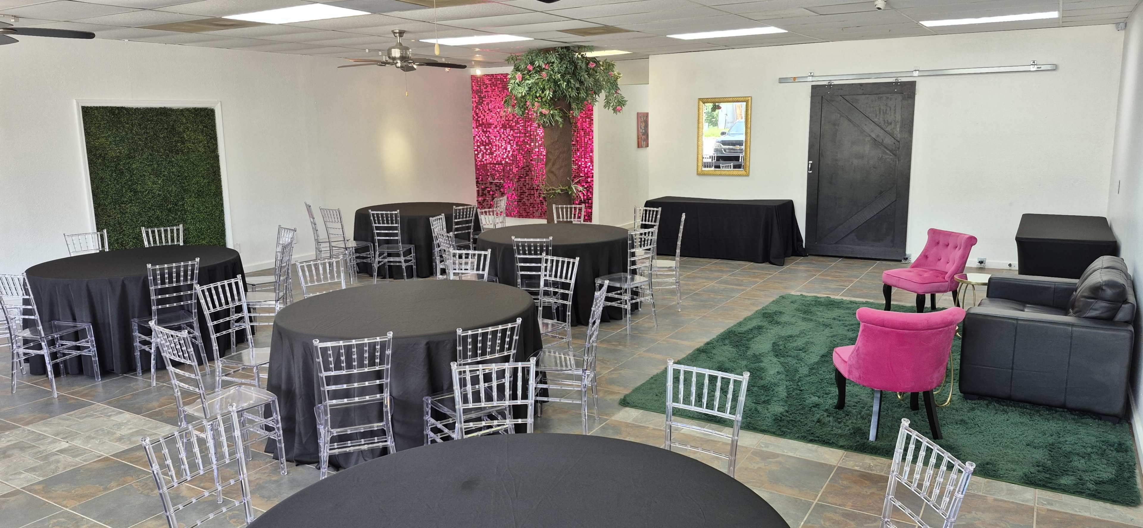 The image shows a spacious event room with multiple round tables covered in black cloth, a pink wall of flowers, and various seating areas including a pink chair and a black couch.
