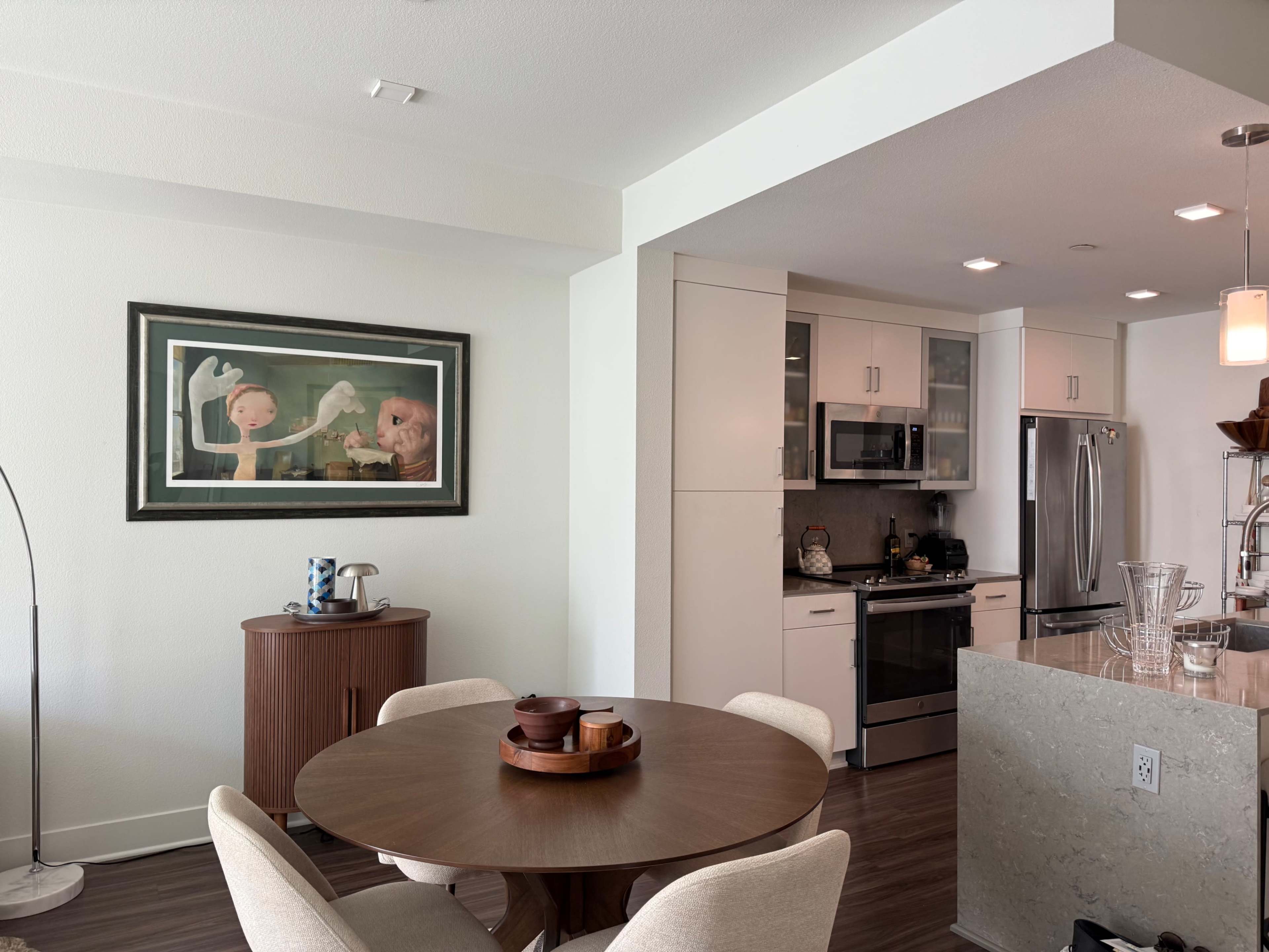 The image shows a modern kitchen and dining area with a circular wooden table, stylish chairs, and framed artwork on the wall.