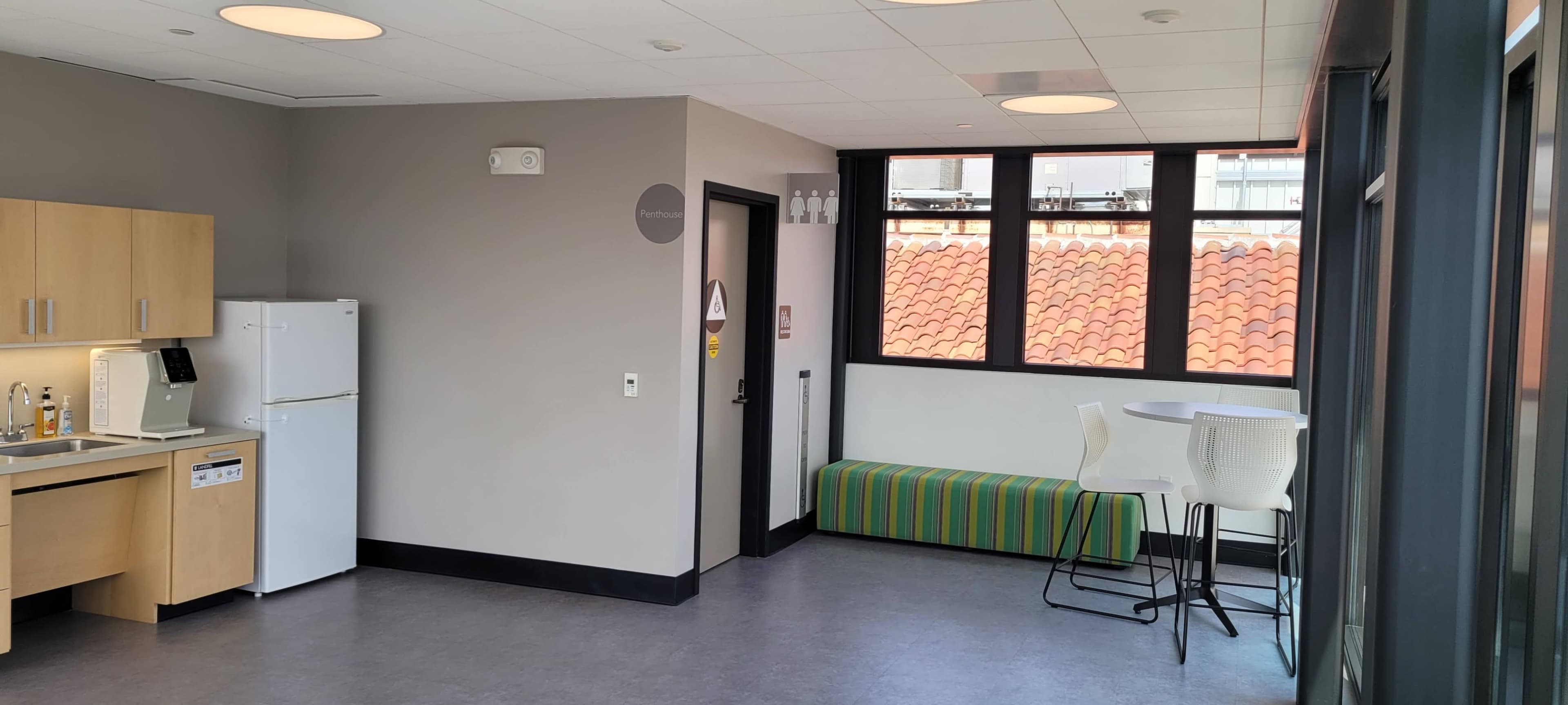 The image shows a modern kitchen area with a refrigerator, sink, and countertop, alongside a seating area with a green-striped bench and high stools, illuminated by natural light from large windows.