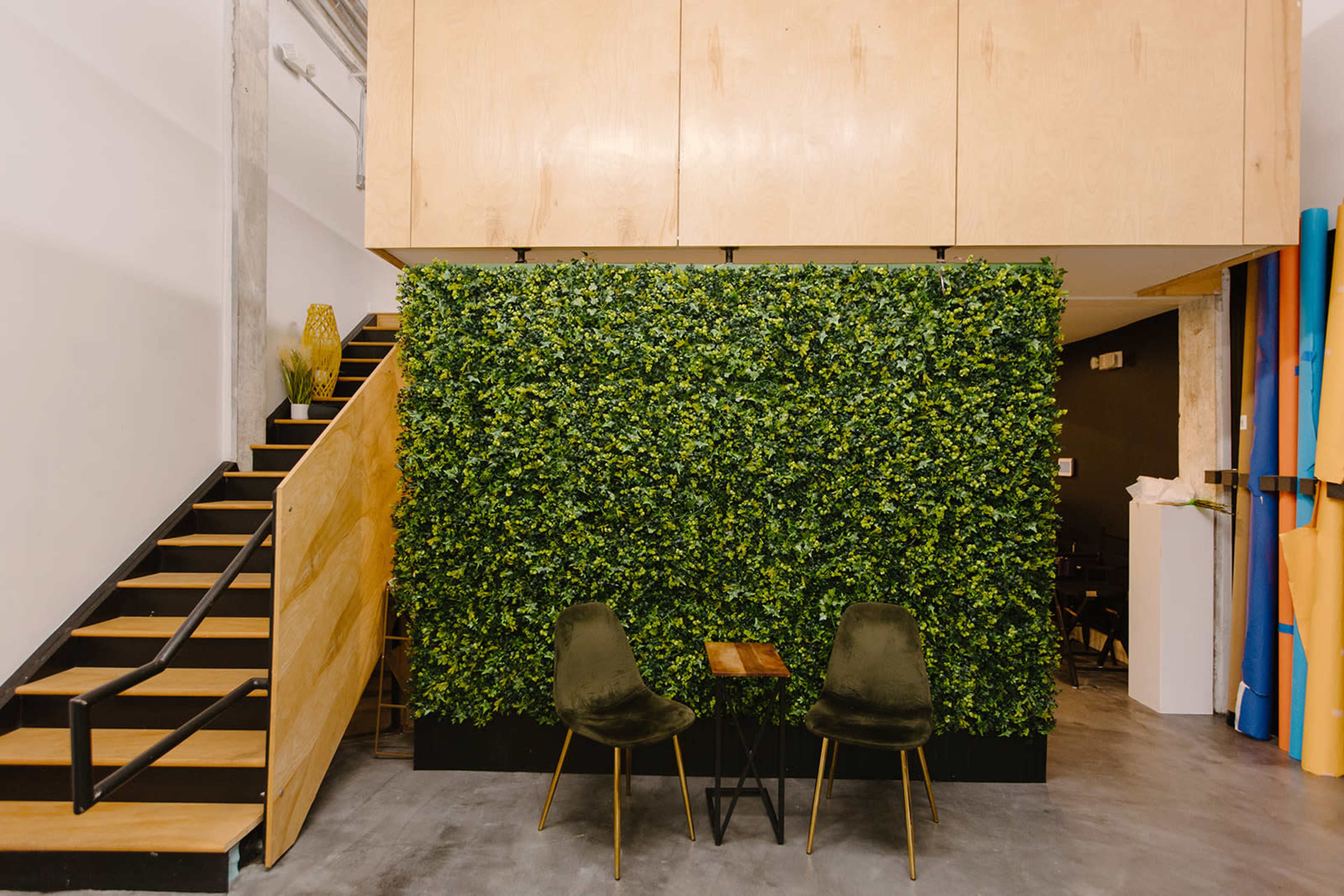 An indoor space featuring a green wall of artificial plants, two chairs, and a small table, with a staircase in the background.