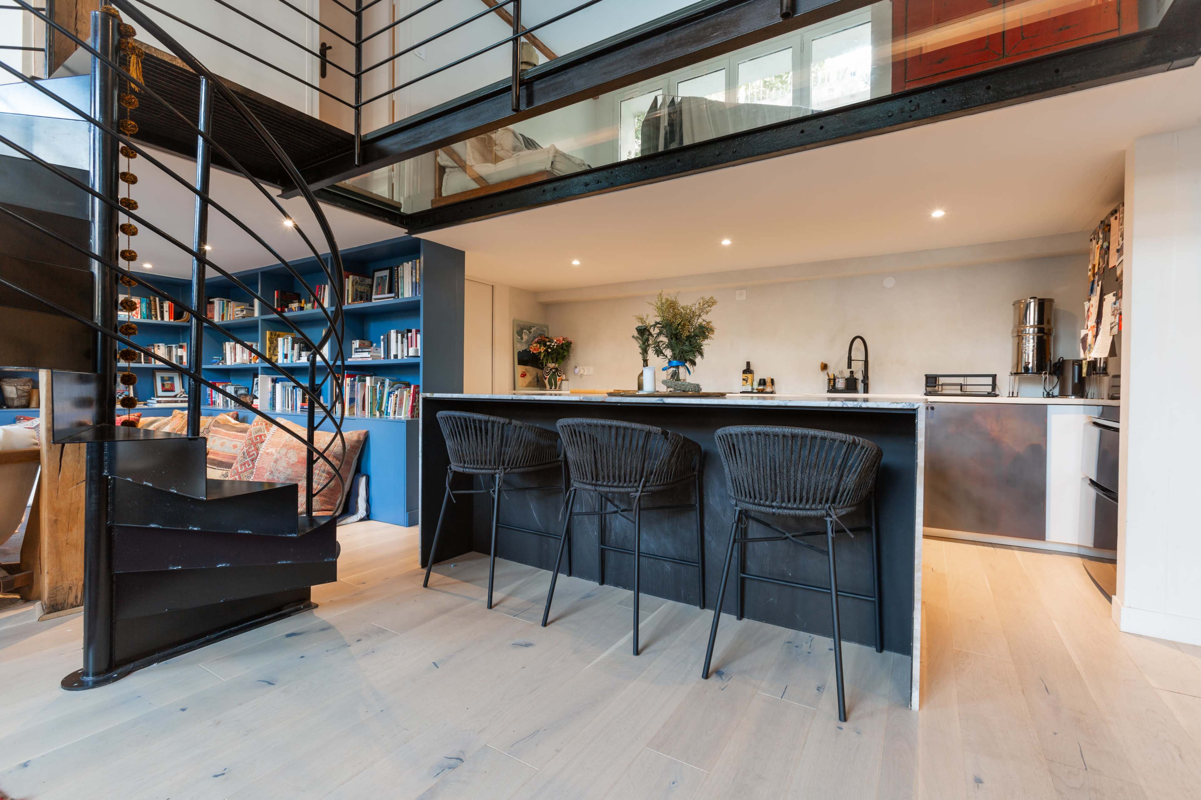 A modern kitchen with a large island and three black bar stools, surrounded by bookshelves and a staircase leading to an upper level.