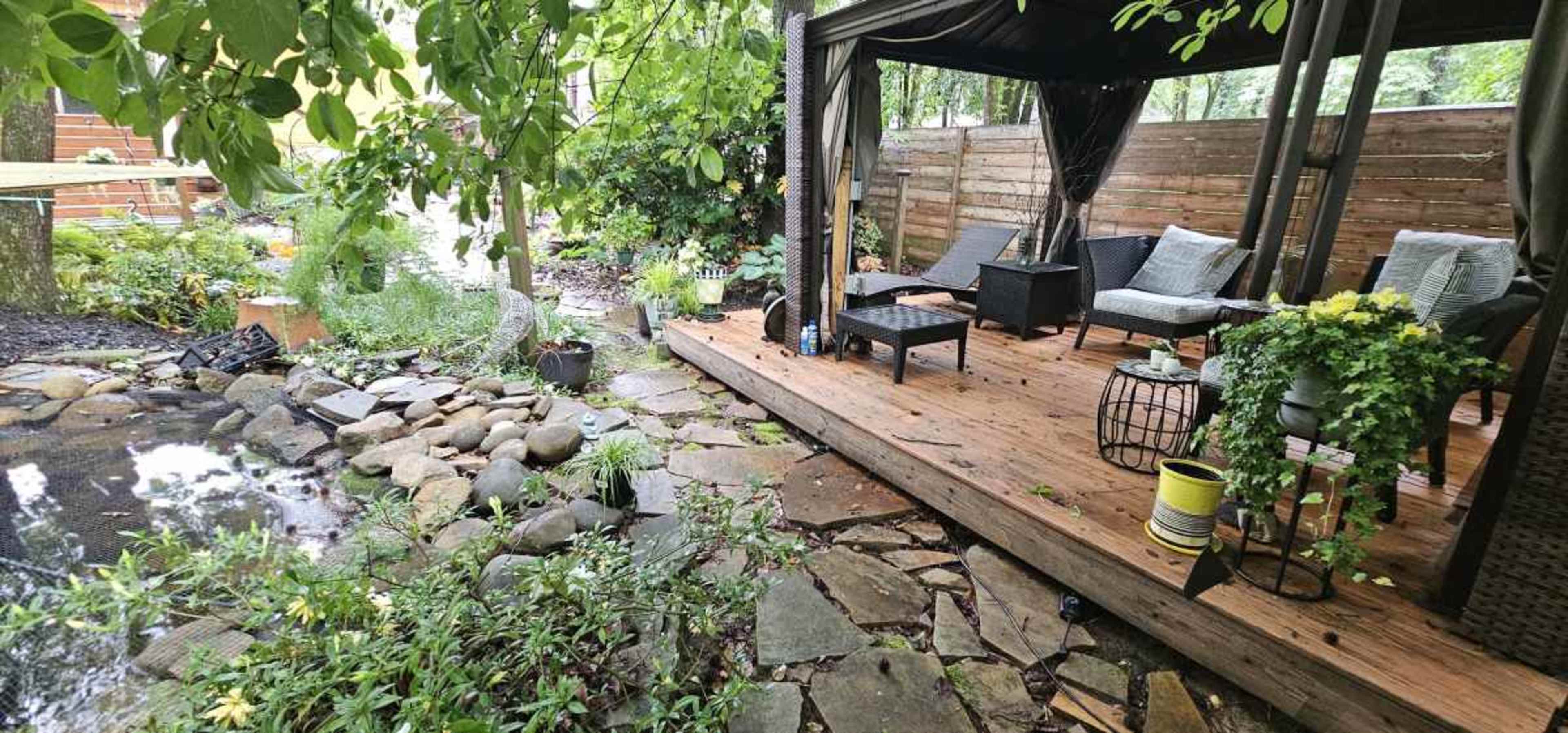 The image shows a wooden patio area with seating and a small pond surrounded by rocks and greenery.