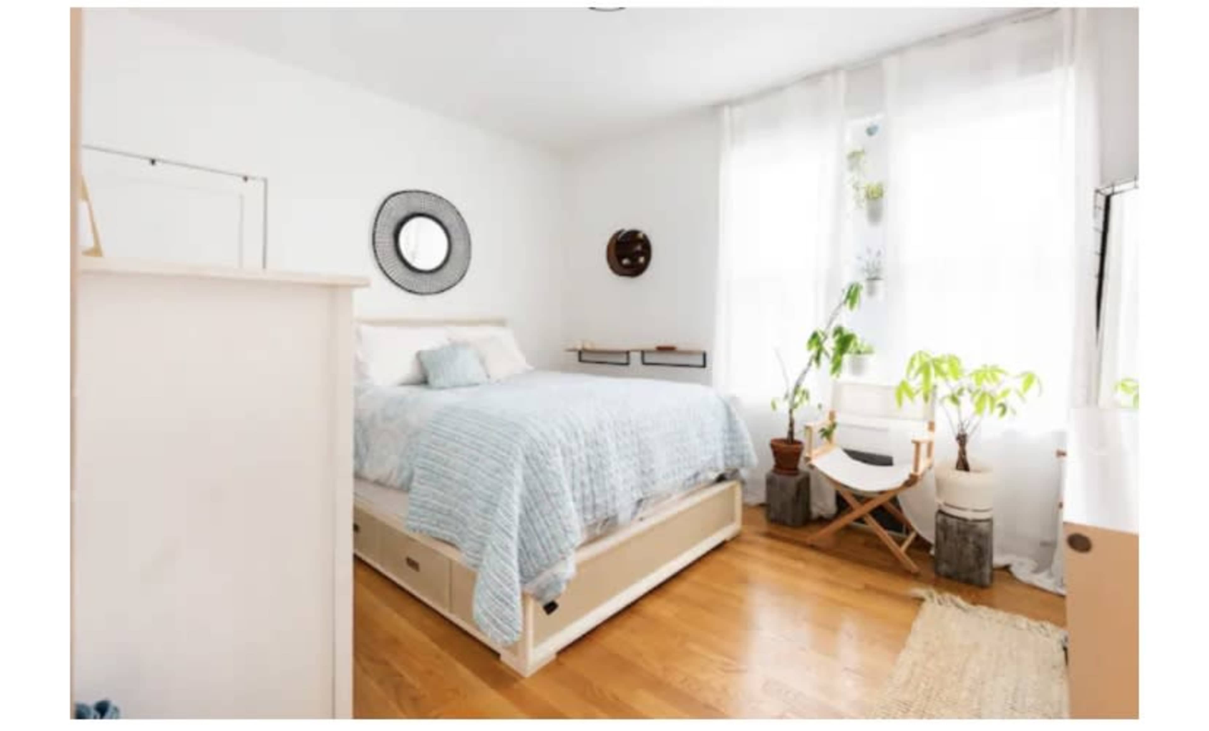 A bright bedroom with a bed, a circular mirror on the wall, a small round table, and plants in natural light.