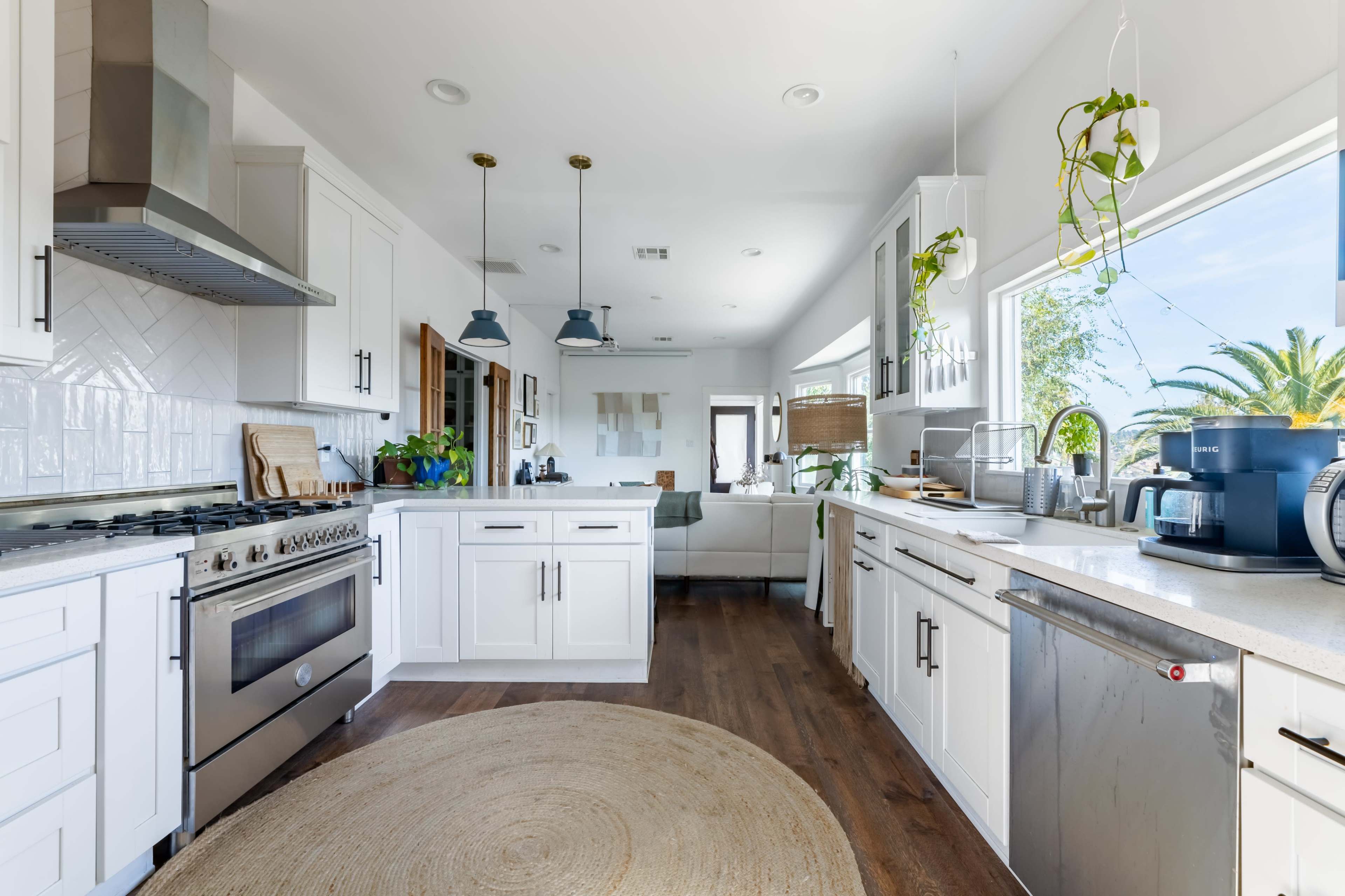 A modern kitchen with white cabinets, stainless steel appliances, and a circular area rug on a wooden floor, leading to a bright living space with plants and large windows.