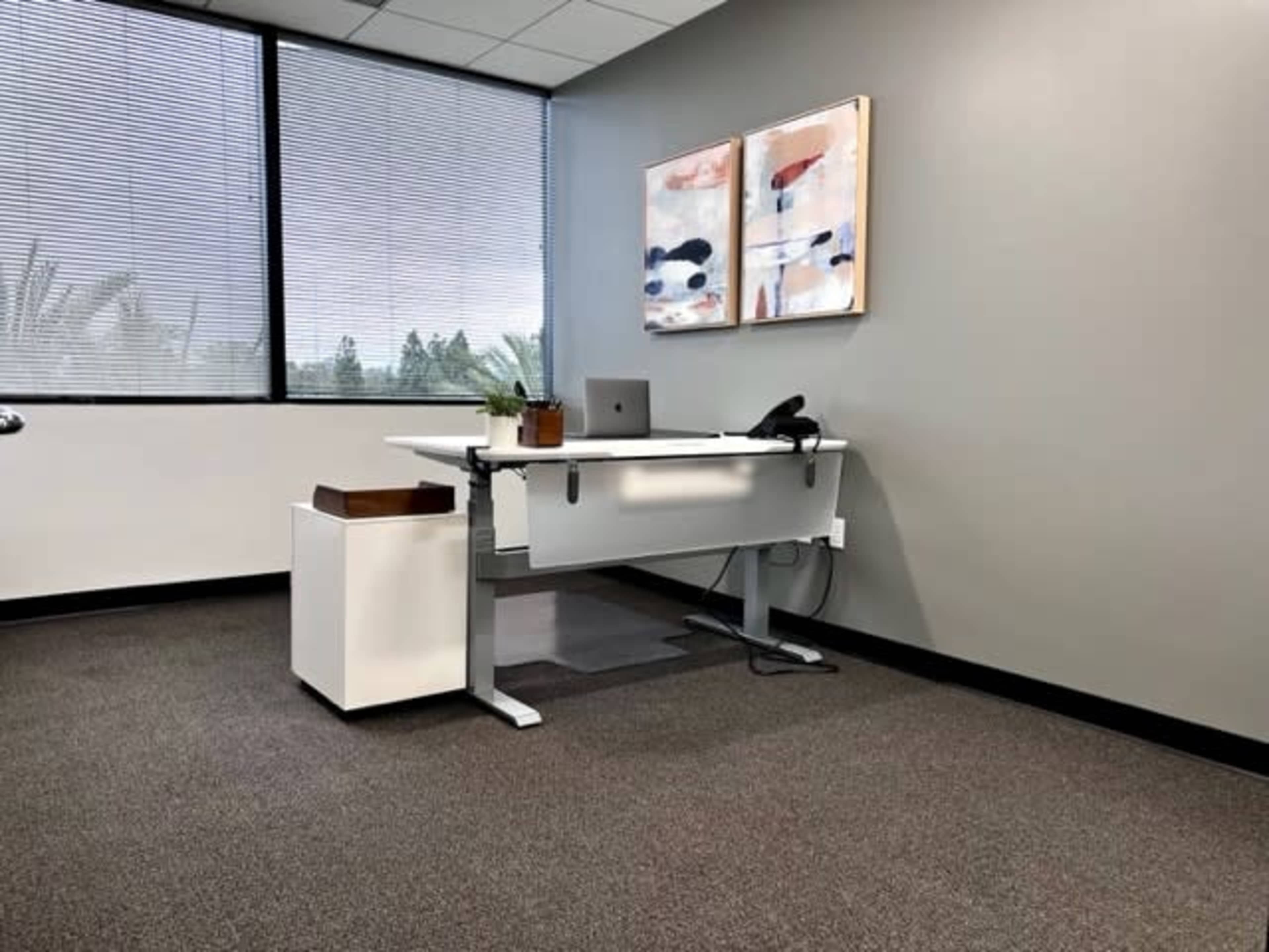 A modern office space featuring a standing desk with a laptop, a phone, and two abstract paintings on the wall.