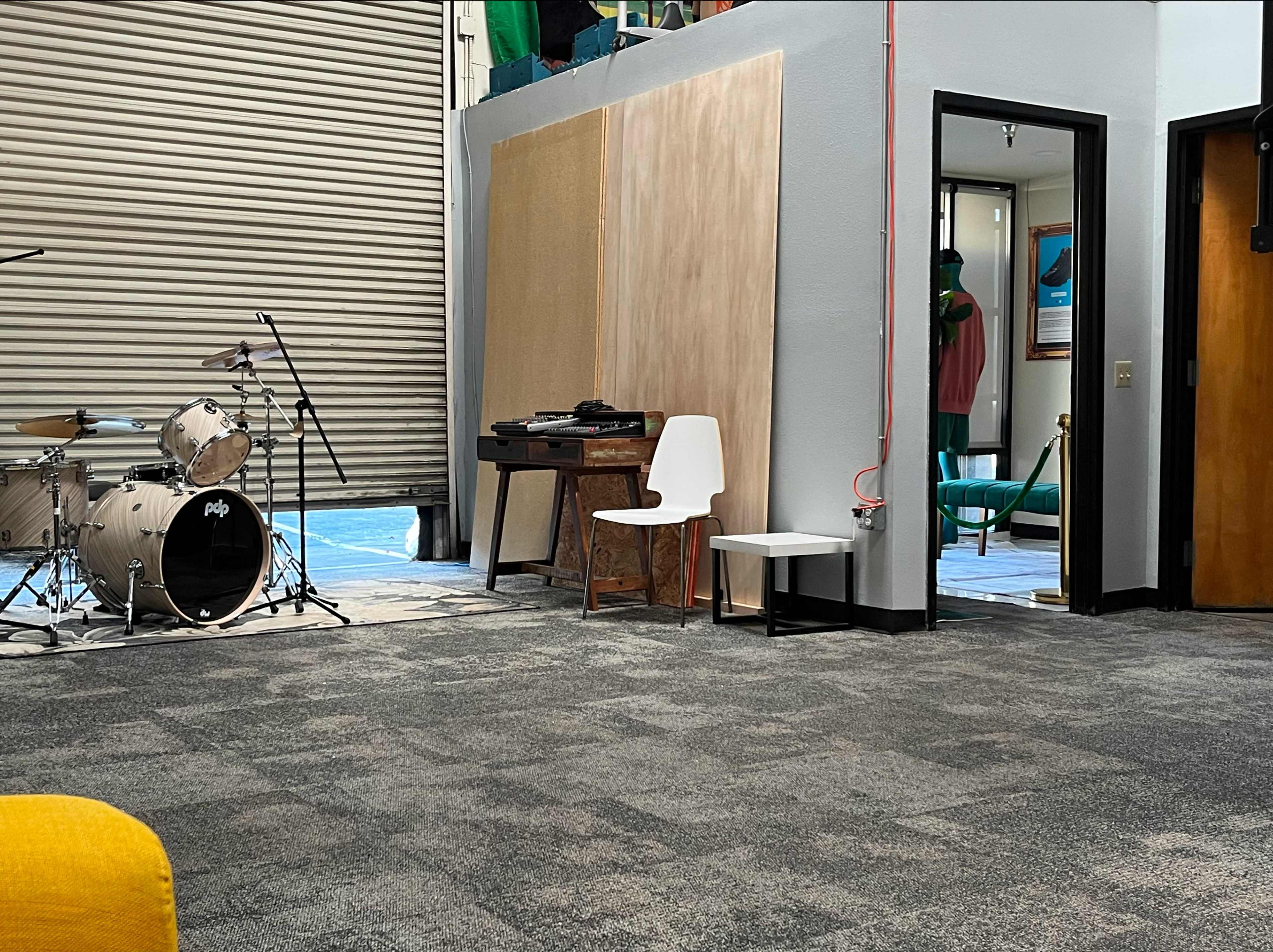 A music setup with a drum kit and electronic keyboard is seen in a spacious room featuring gray carpet and a large garage door on one side.