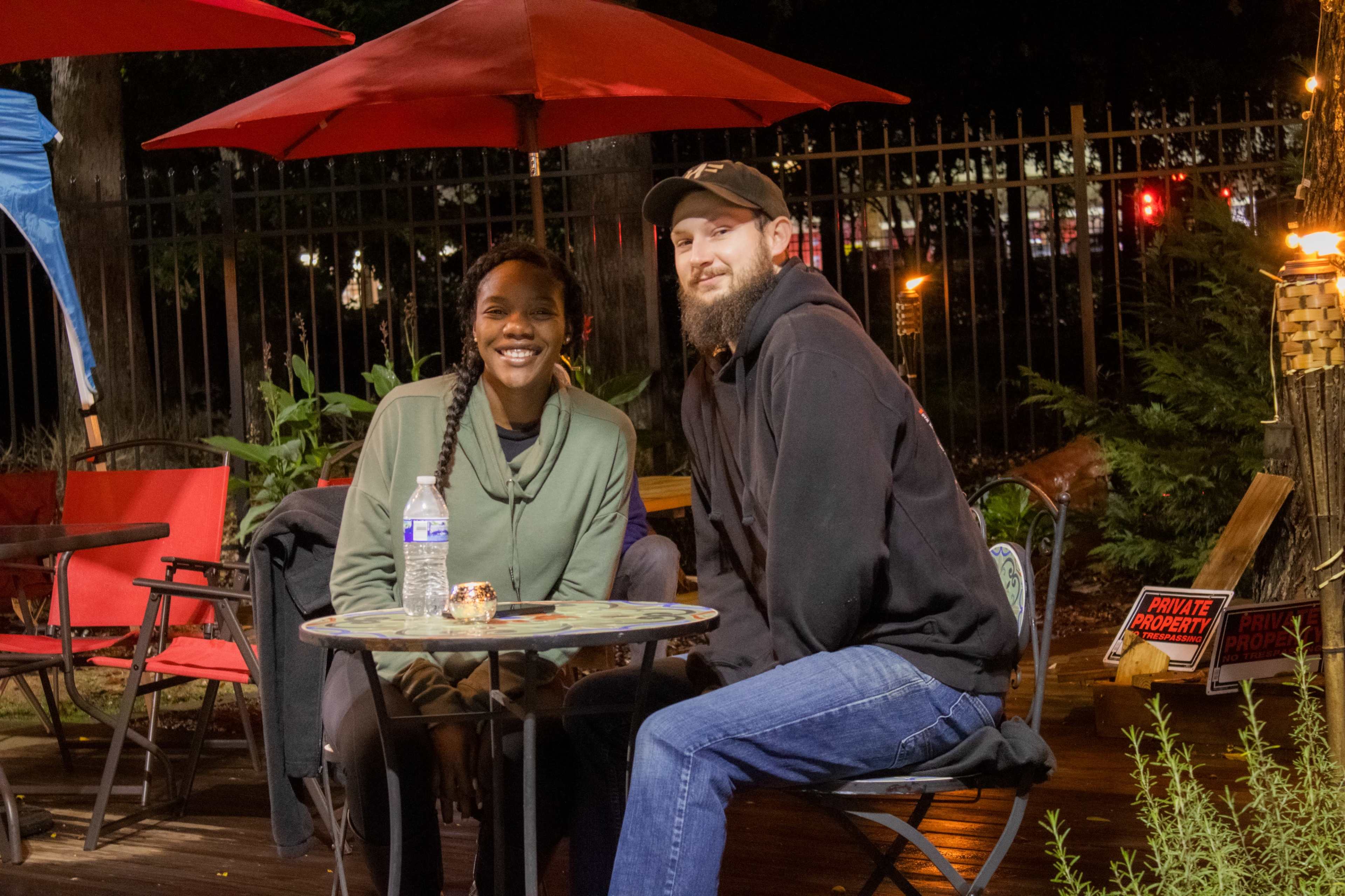 A man and a woman are sitting at a small table with a drink, under red umbrellas in an outdoor seating area.