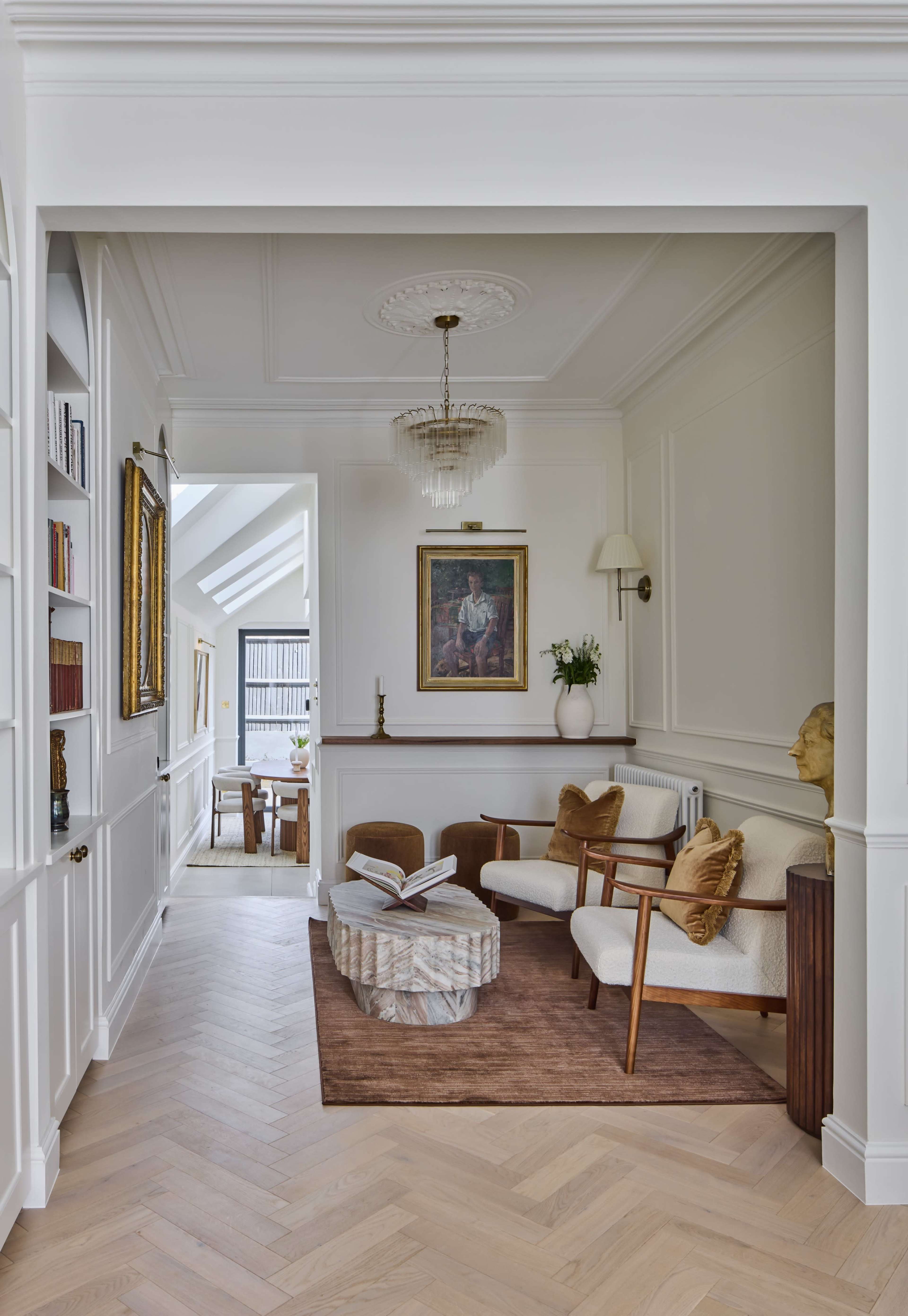 The image shows a neatly designed interior hallway featuring two armchairs, a small table, a chandelier, and framed artwork, with wooden flooring and light-colored walls.