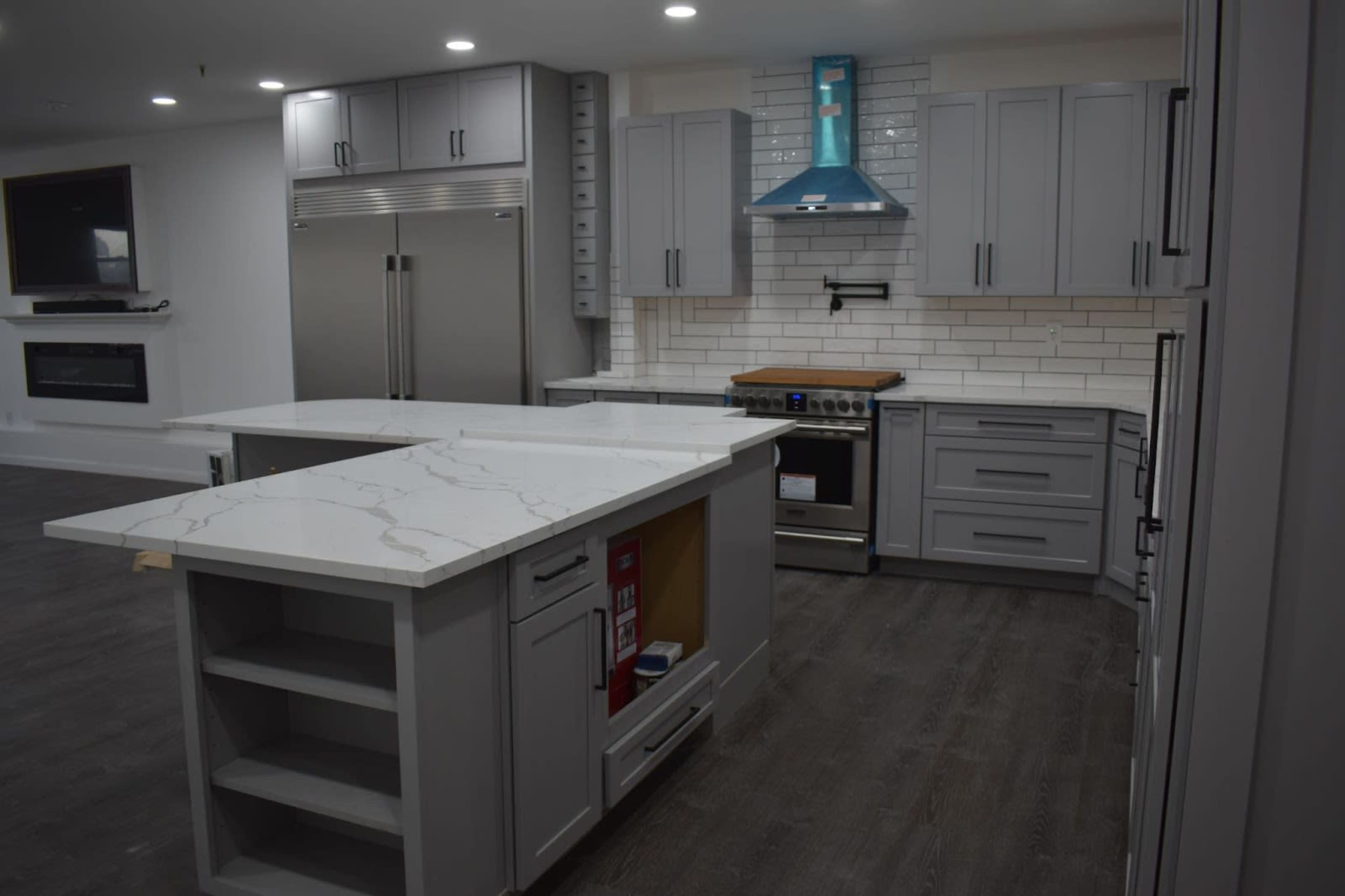 A modern kitchen with gray cabinetry, a large white marble island, and stainless steel appliances, including a range and refrigerator.