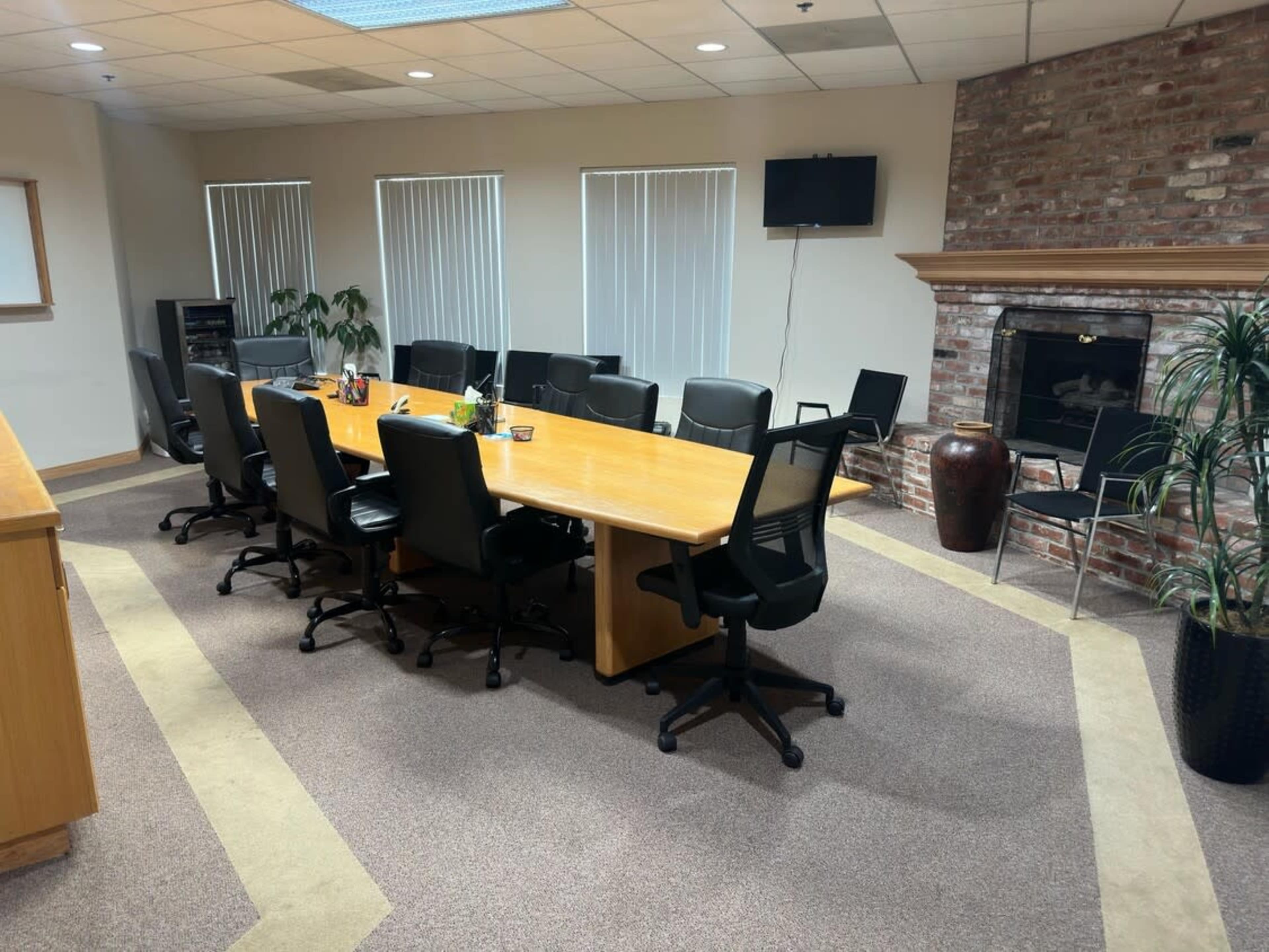A conference room features a large wooden table surrounded by swivel chairs, with a brick fireplace and a TV mounted on the wall.