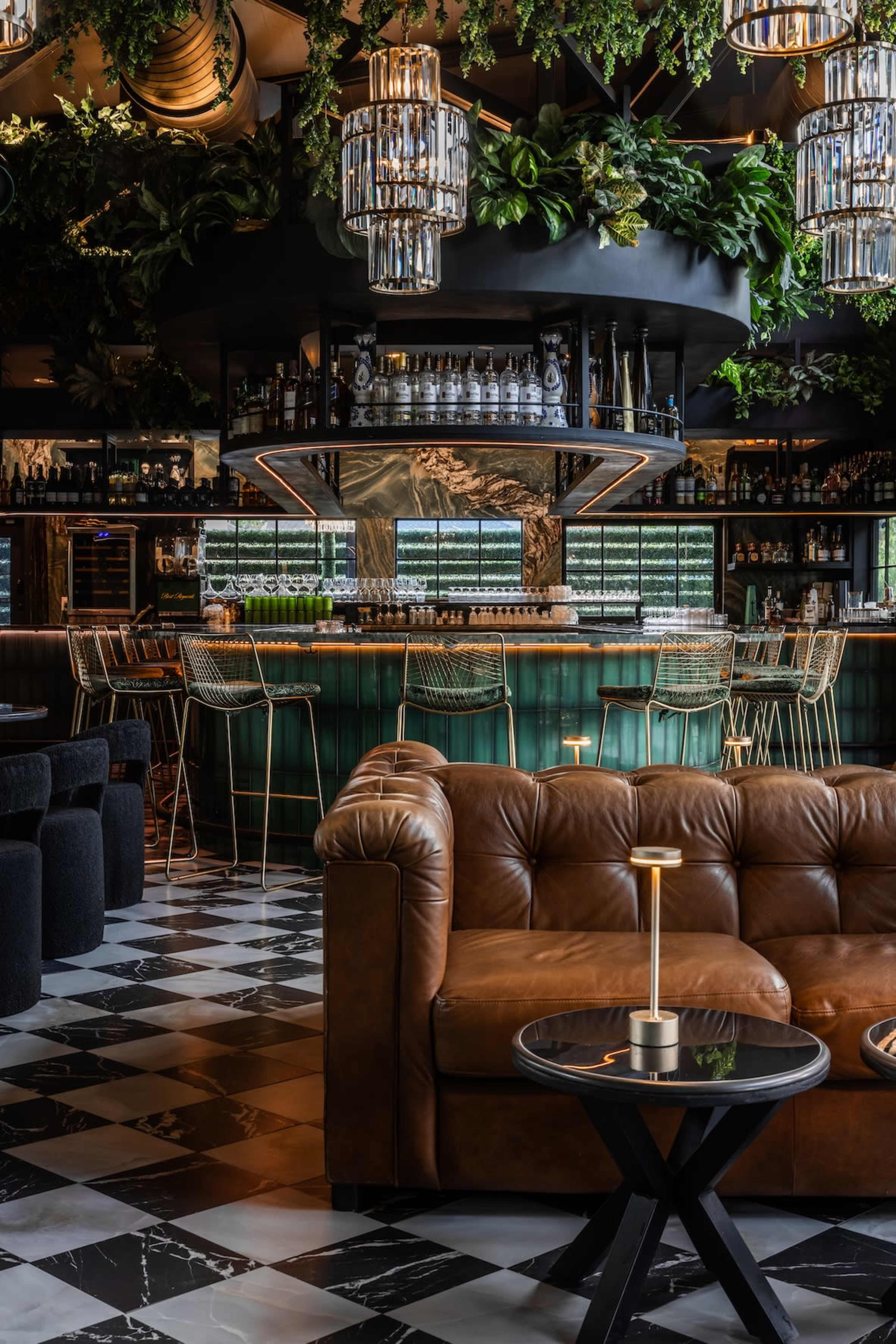 The image shows a stylish bar area with a green and black color scheme, featuring plush seating, intricate light fixtures, and a well-stocked bar surrounded by greenery.