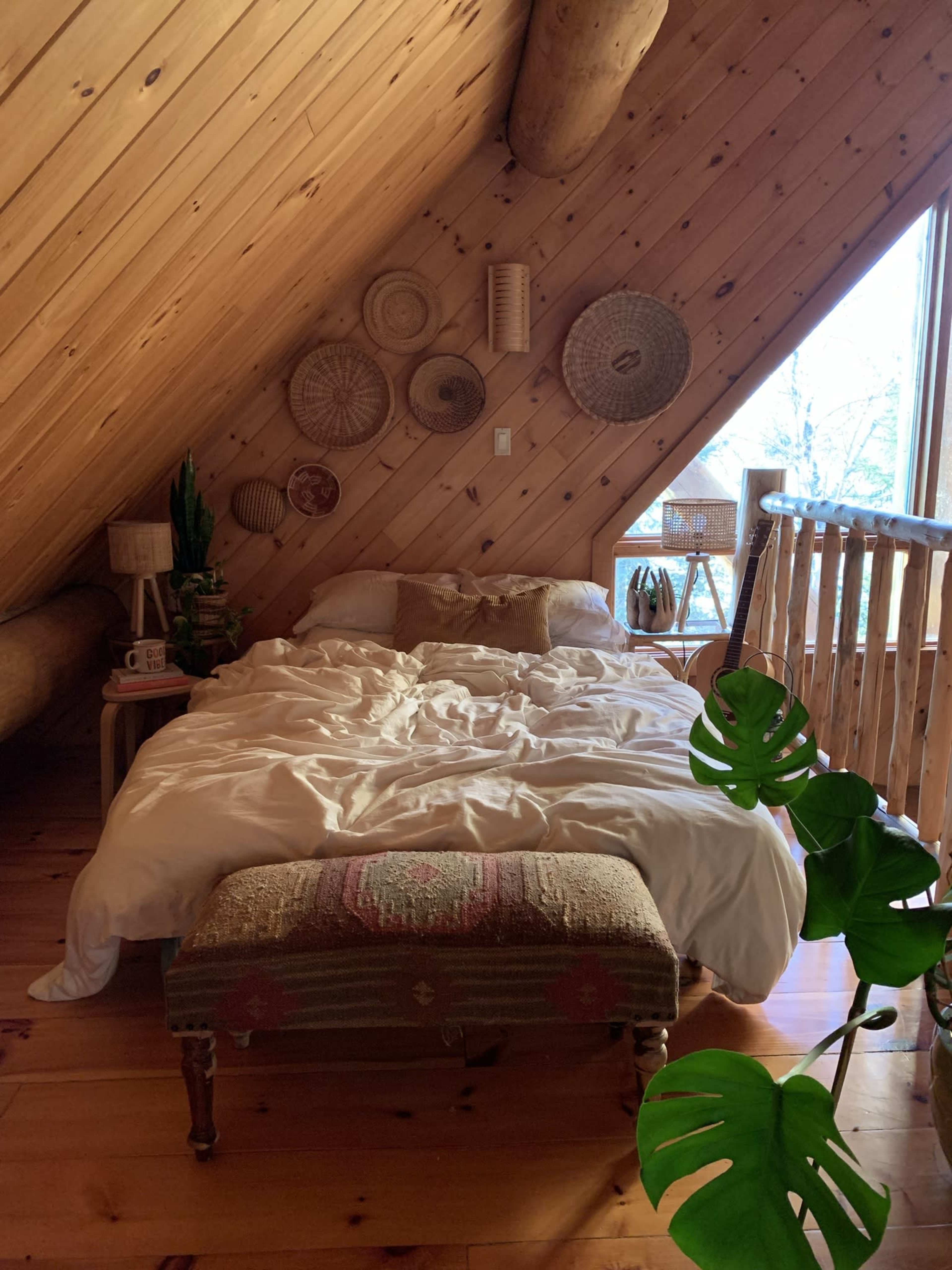 The image shows a cozy attic bedroom with a bed covered in white bedding, a bench at its foot, and decorative wall baskets, all illuminated by natural light from a large window.