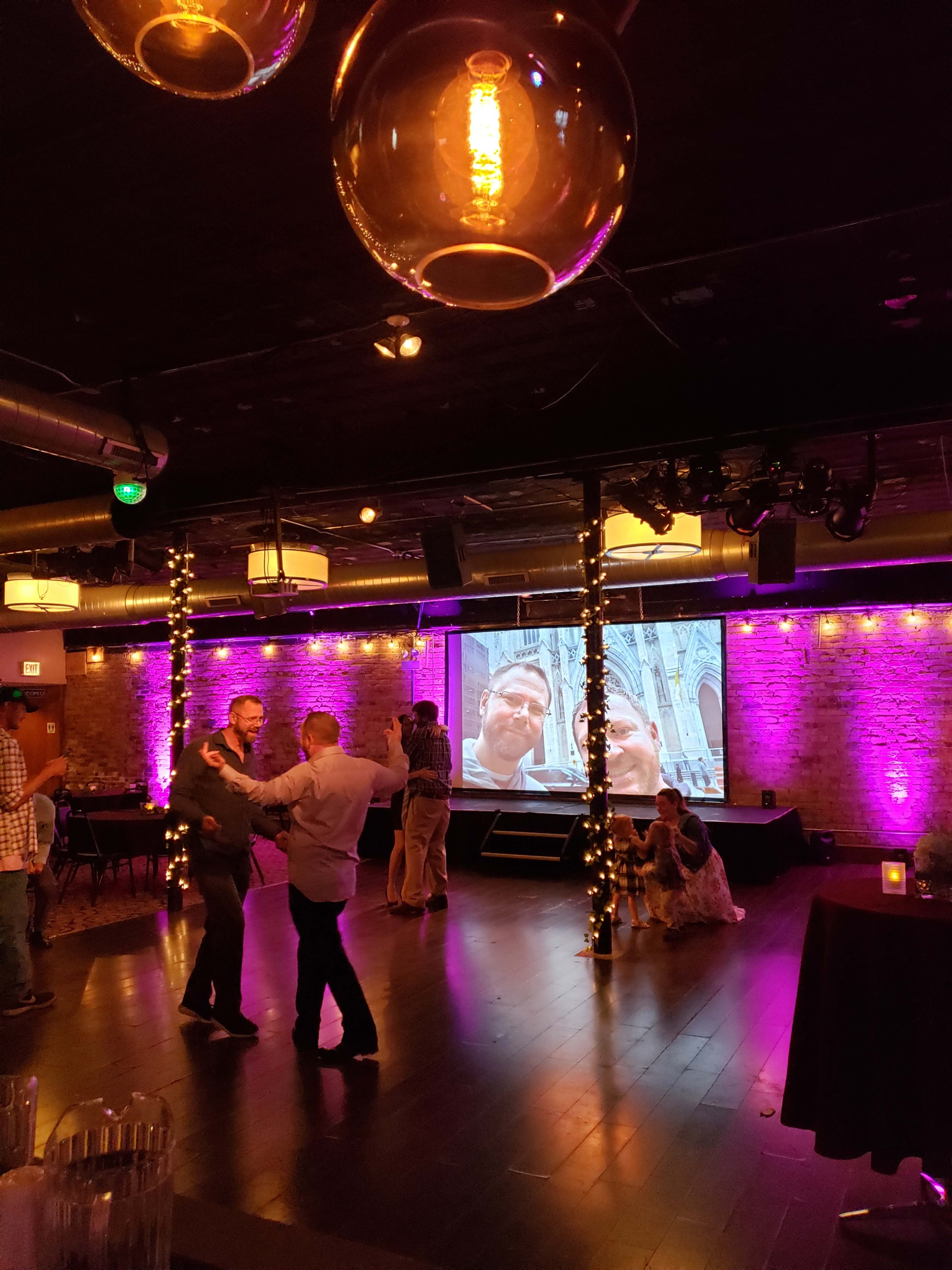 People are dancing in a dimly lit venue with brick walls and a large screen displaying a photo in the background.