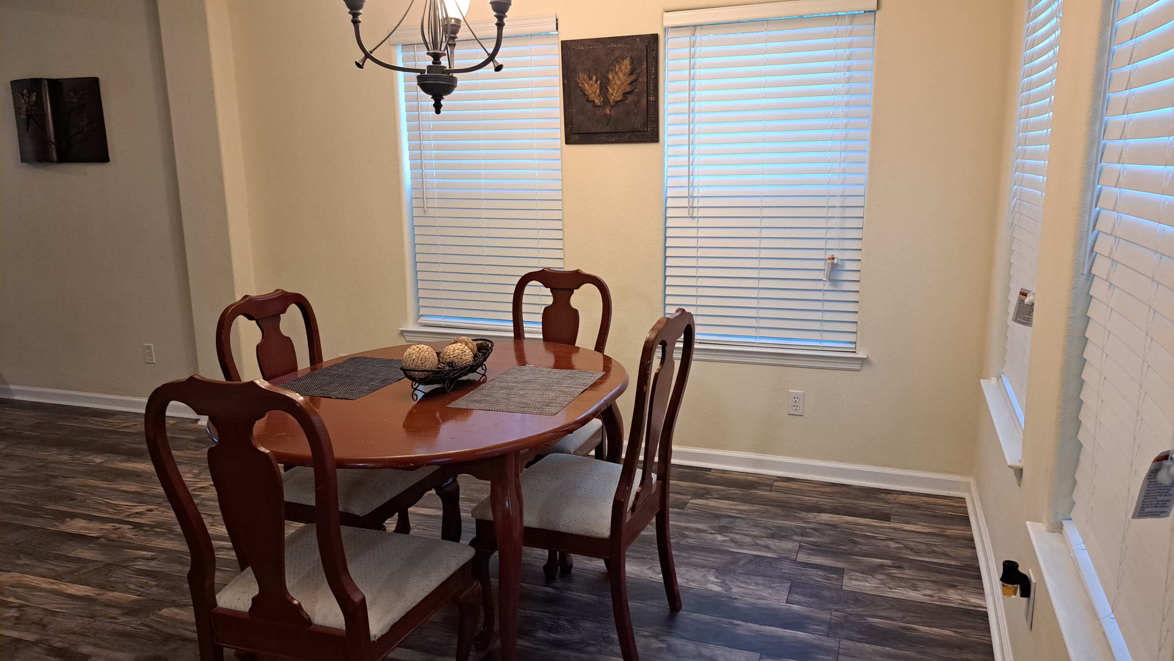 A round wooden dining table is set with four chairs in a room featuring two windows with blinds.
